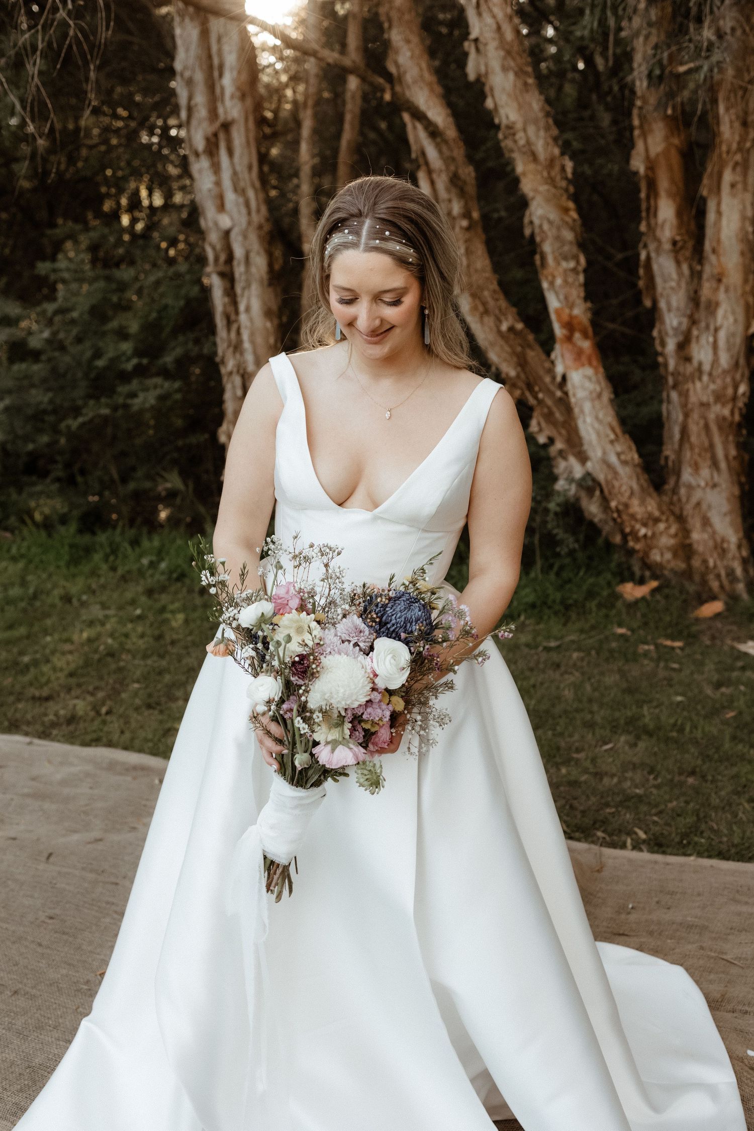 Bride in deep V-neck white ball gown admiring colorful floral bouquet in a wooded outdoor setting.