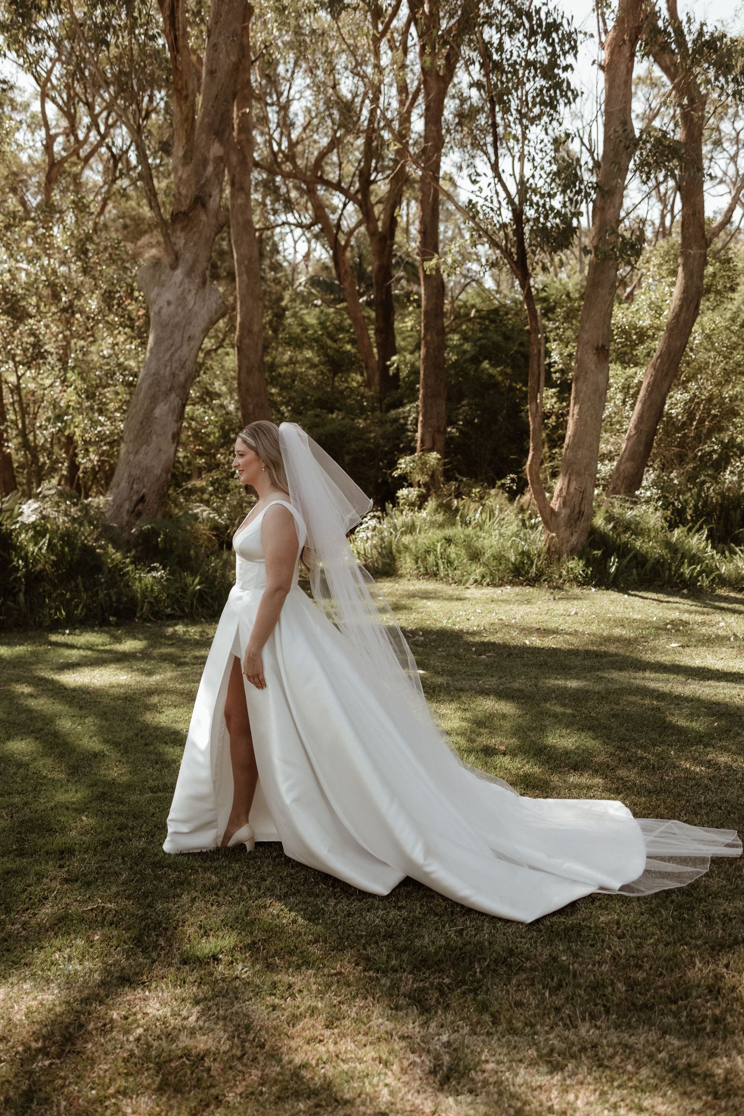 Elegant bride in a white gown with long veil walking through a sunlit forest on her wedding day.