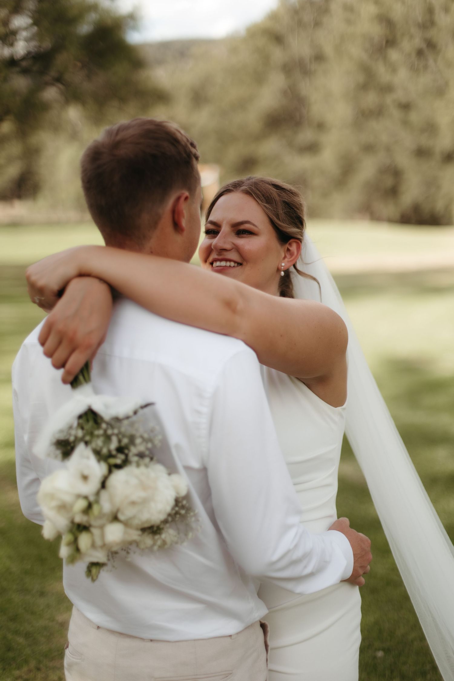 Radiant bride hugs groom while holding white bouquet on a lush green lawn.