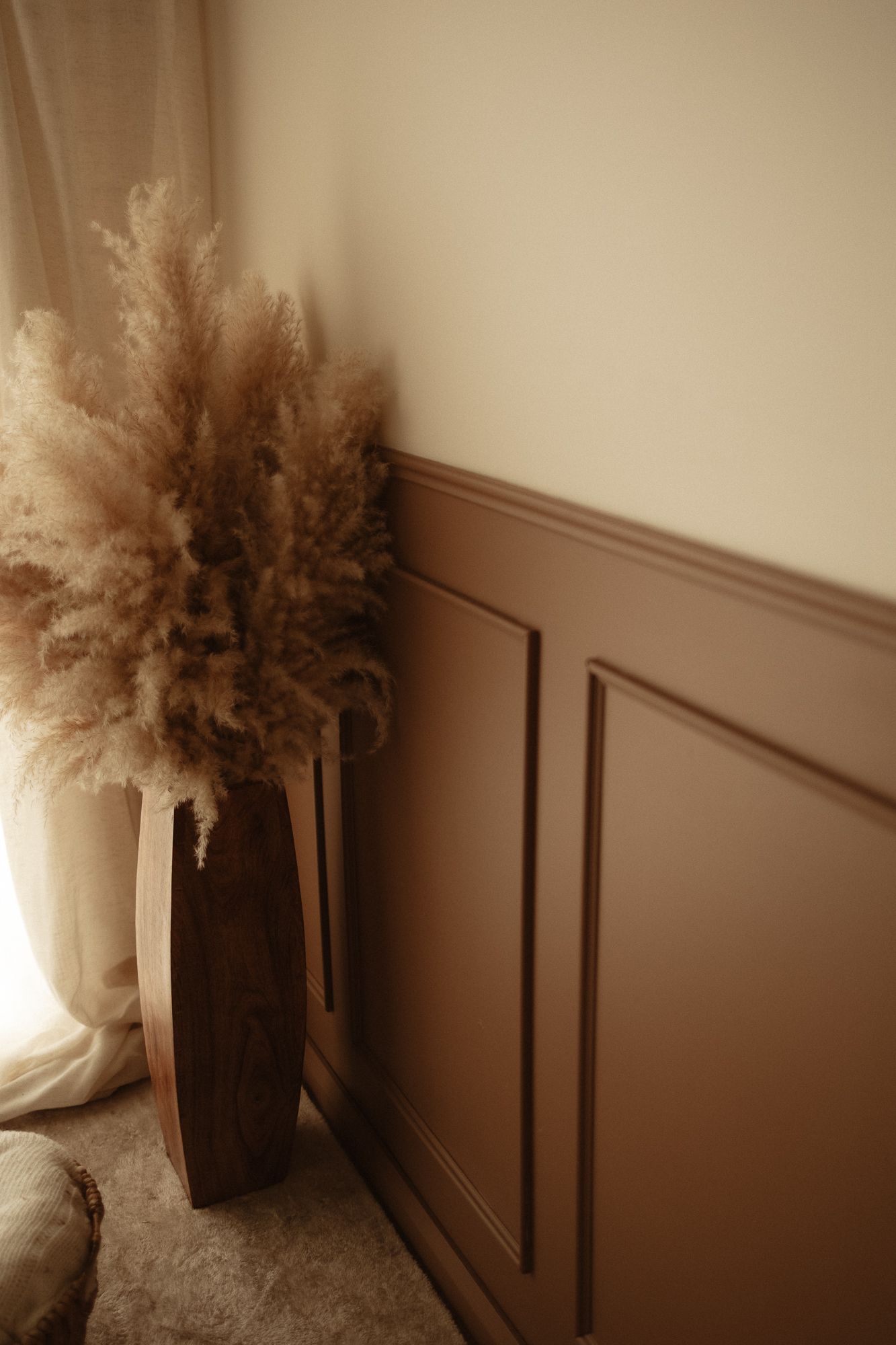 Brown wainscoting wall panel with dried pampas grass in a wooden vase near a sheer curtain window.