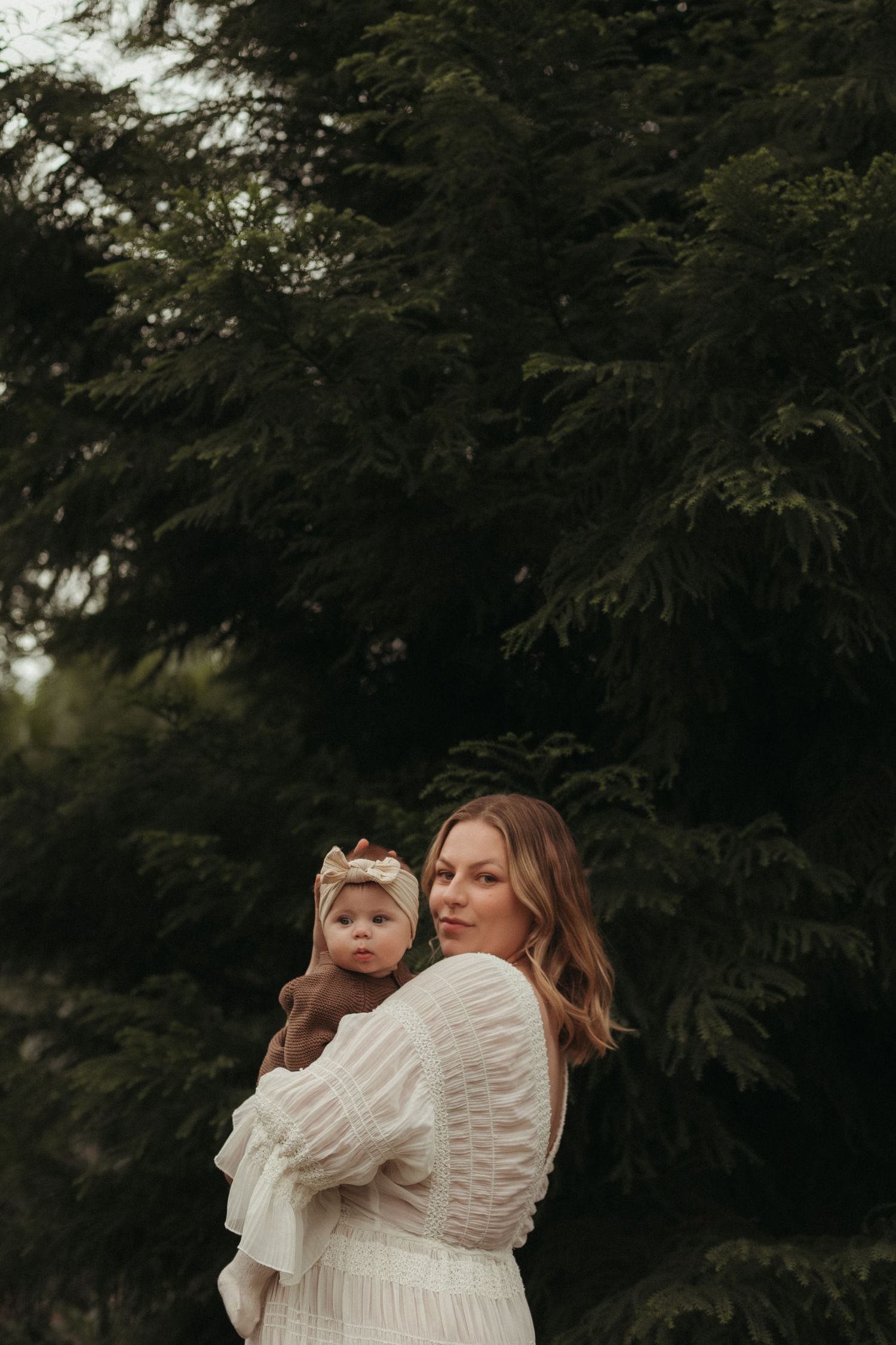 Mother in white knit dress holding baby with floral headband outdoors.