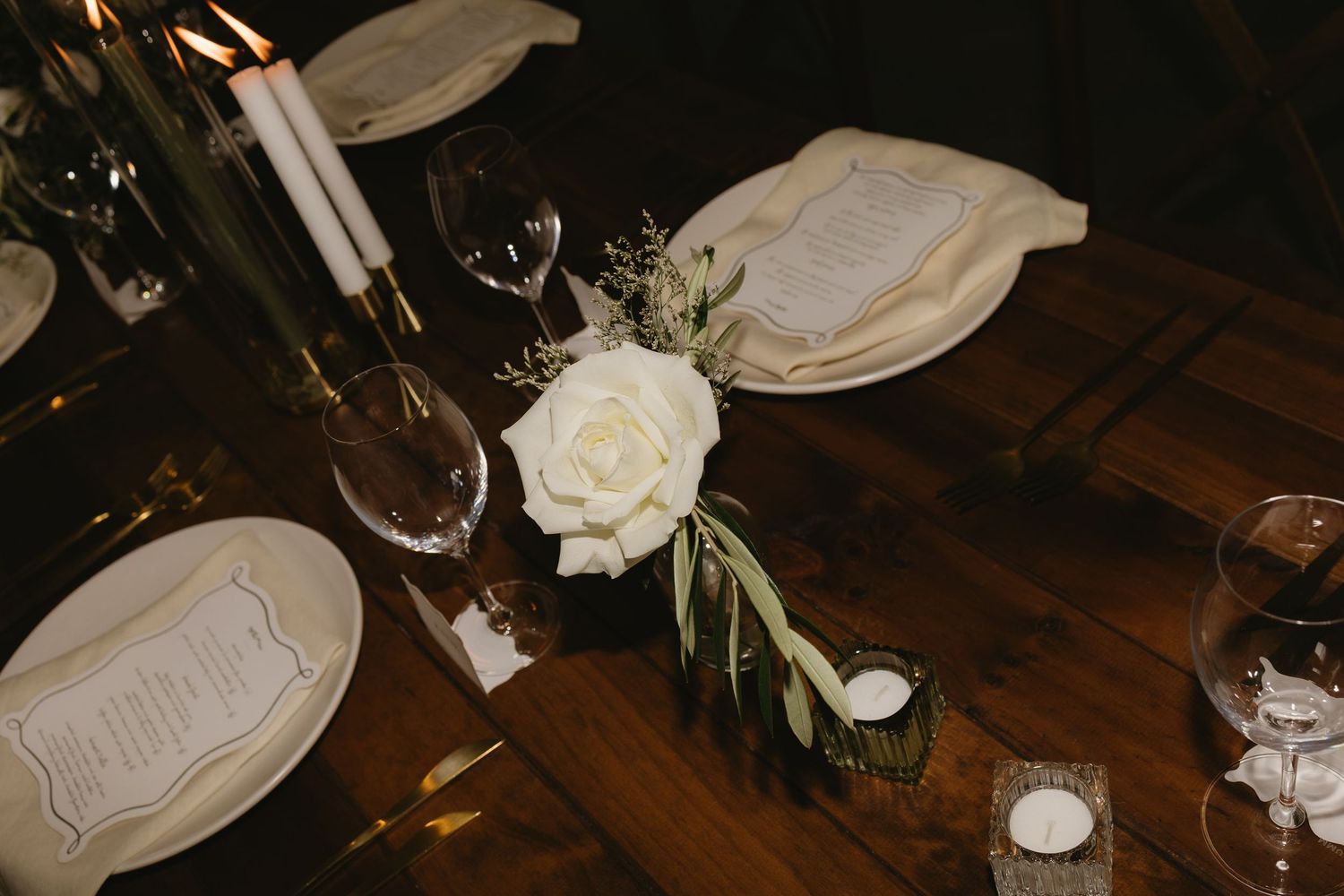 Close-up of a wedding table setting featuring a white rose, menu cards, candles, and glassware.