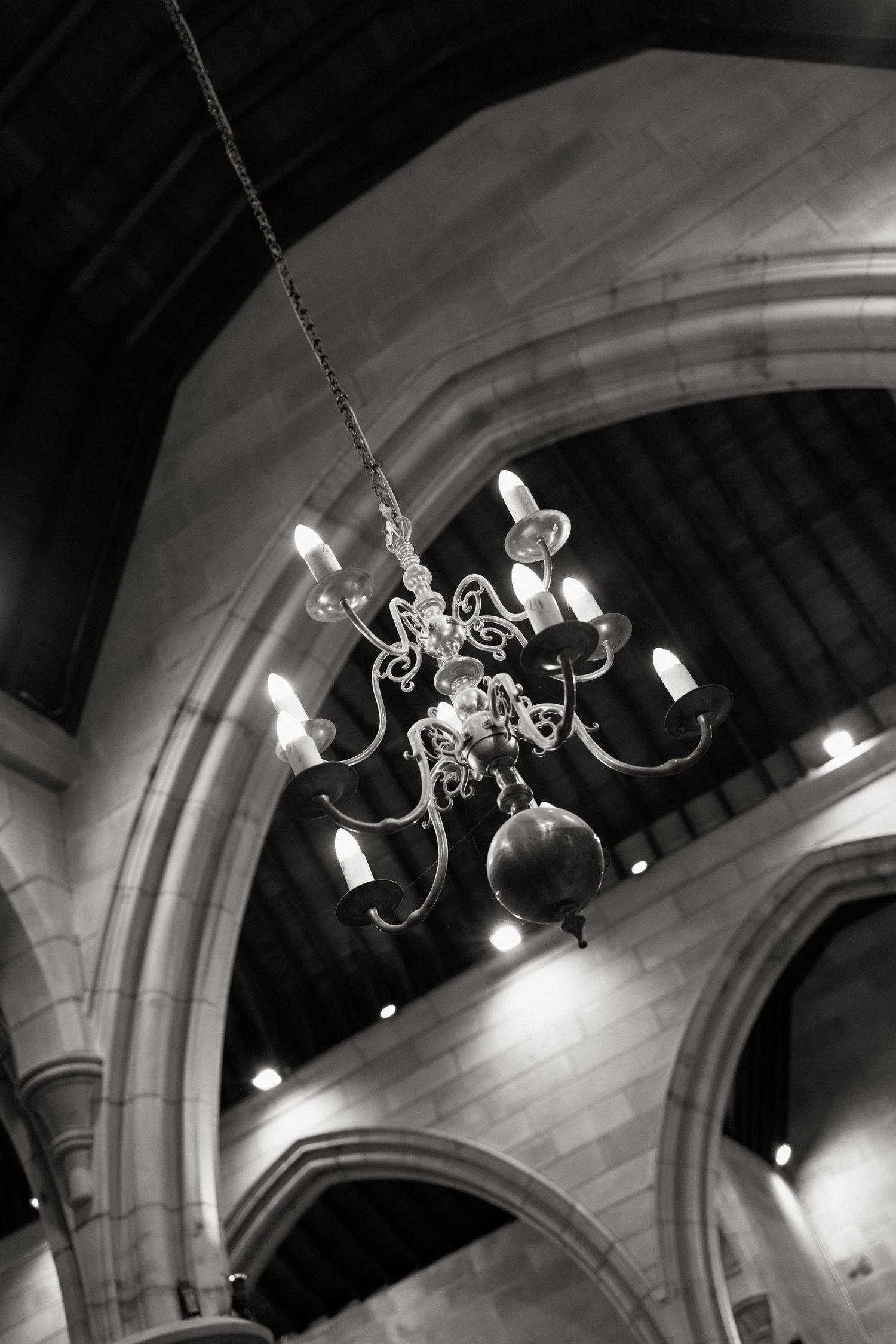 Black and white photo of an ornate chandelier hanging beneath a vaulted stone arch in a historic church.