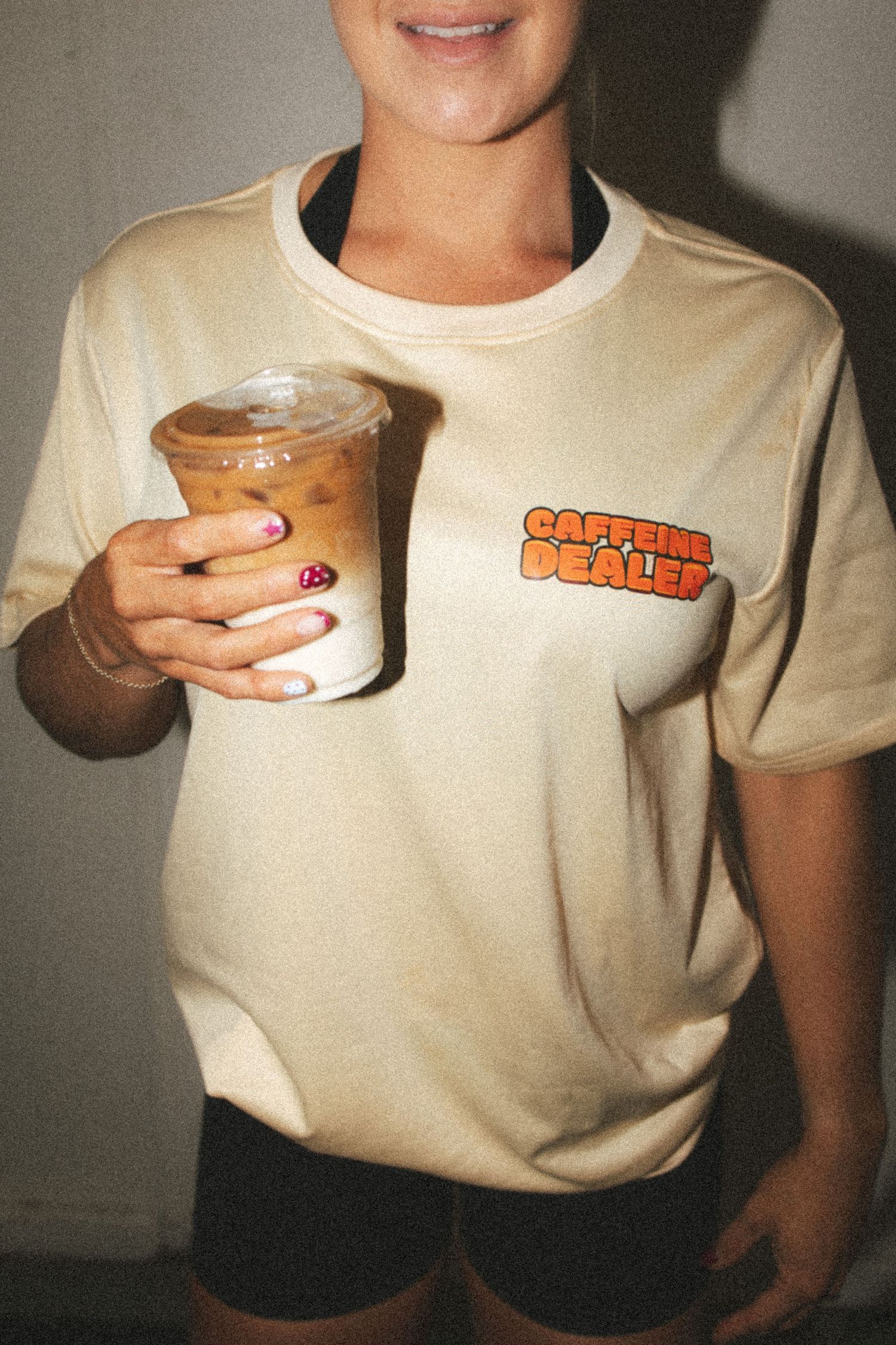 Woman in cream Caffeine Dealer t-shirt holding an iced coffee drink with pink manicured nails.