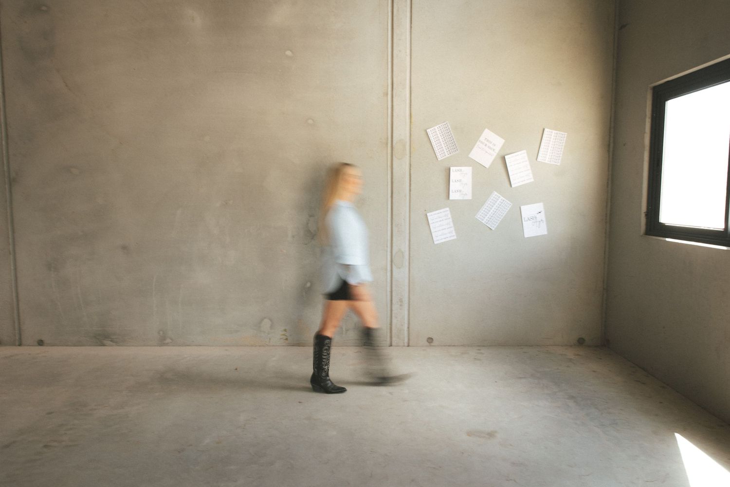 Blurred woman walking past concrete wall with papers pinned to it in empty industrial space.