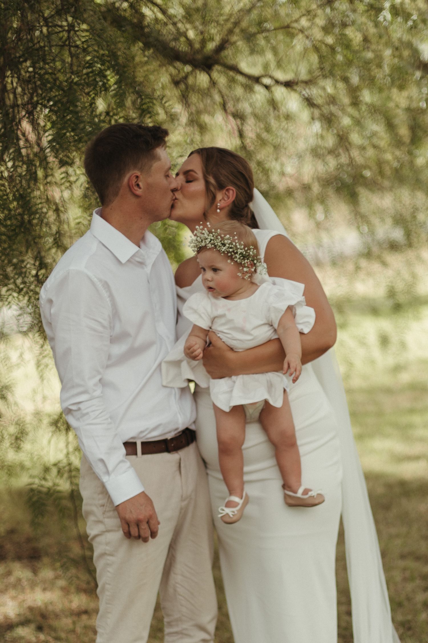 A couple in white wedding attire kisses while holding their baby girl in a floral crown outdoors.