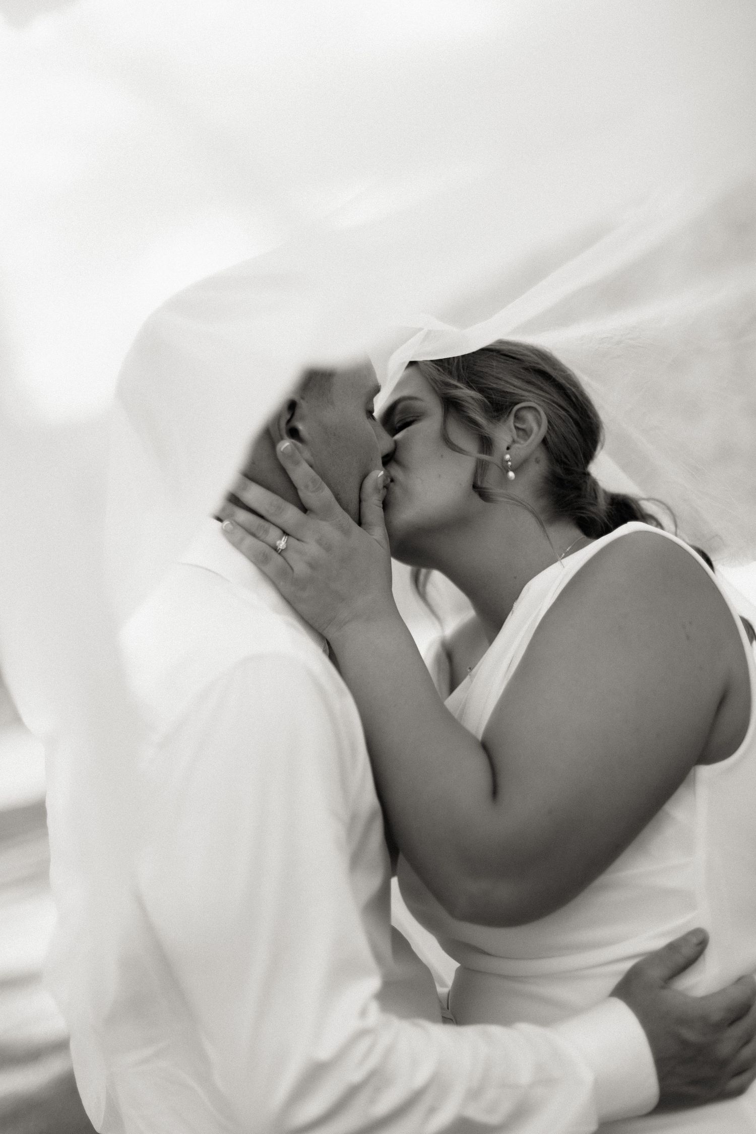 Black and white photo of a bride and groom sharing a romantic kiss under a flowing wedding veil.