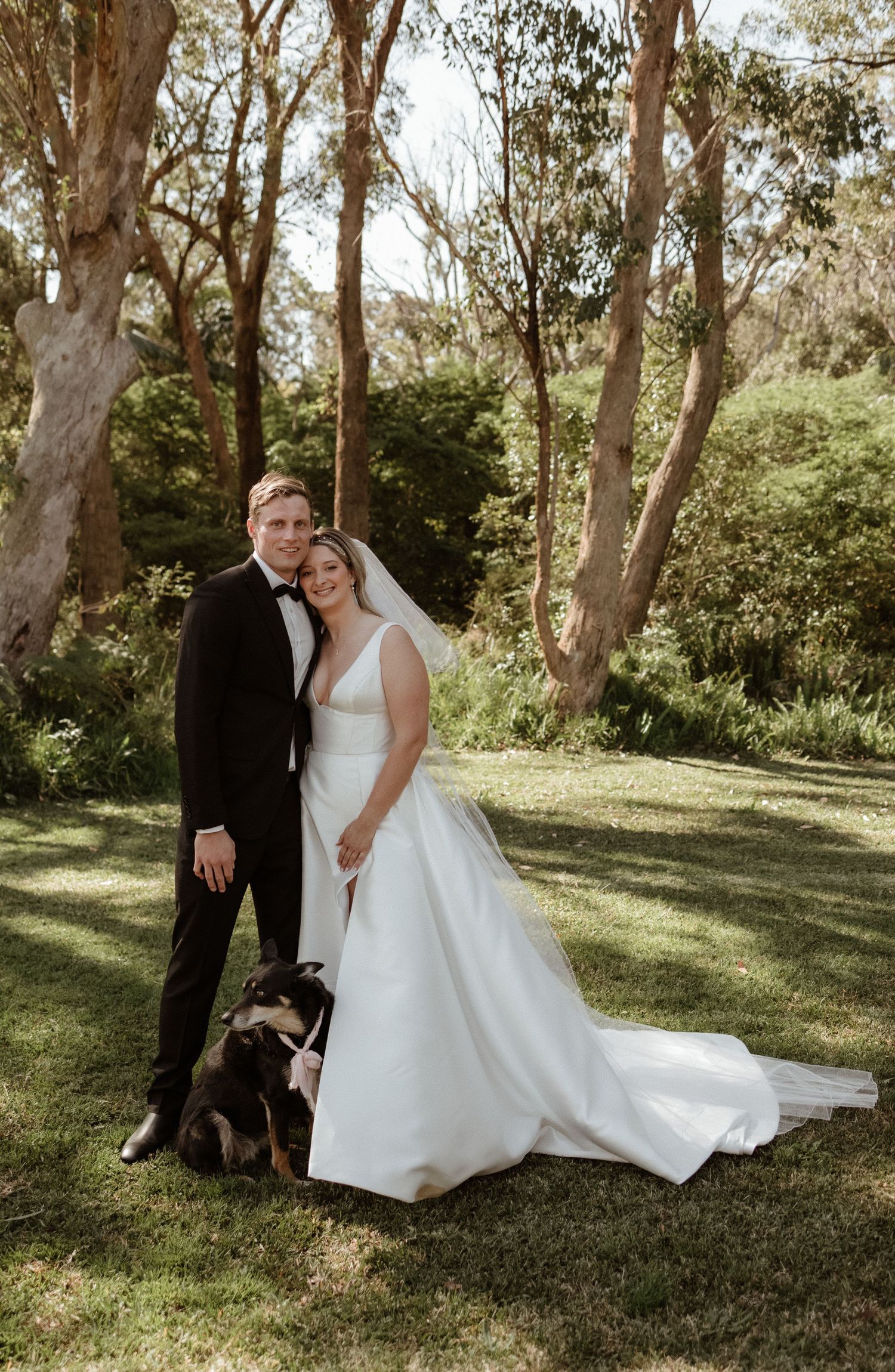 Bride and groom pose with their dog in a lush garden setting, bride in white gown, groom in black tuxedo.