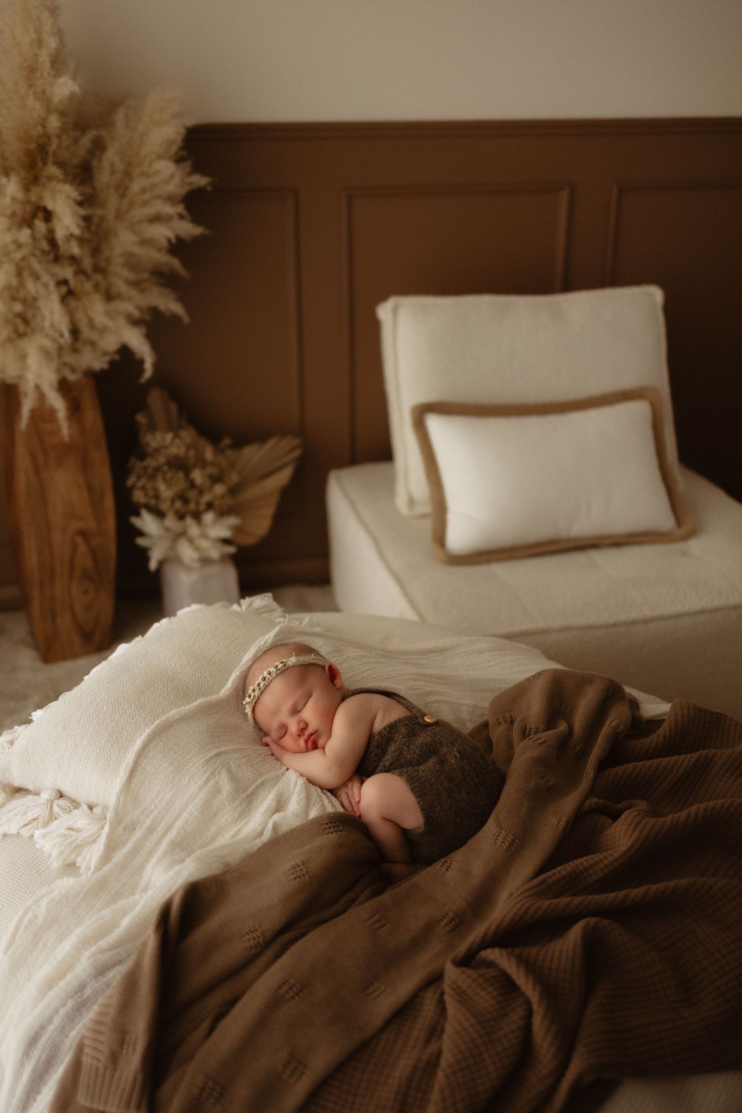 Newborn baby sleeping peacefully on a brown blanket in a boho-styled bedroom with pampas grass decor.