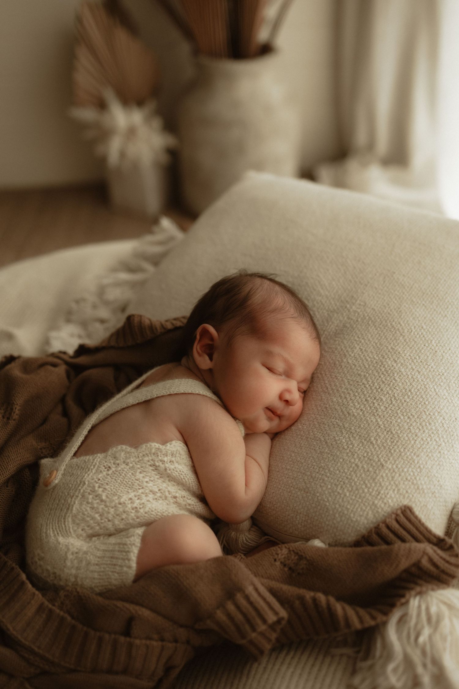 Peaceful newborn baby sleeping on a cream knit pillow wrapped in a brown blanket, wearing white romper in boho nursery.