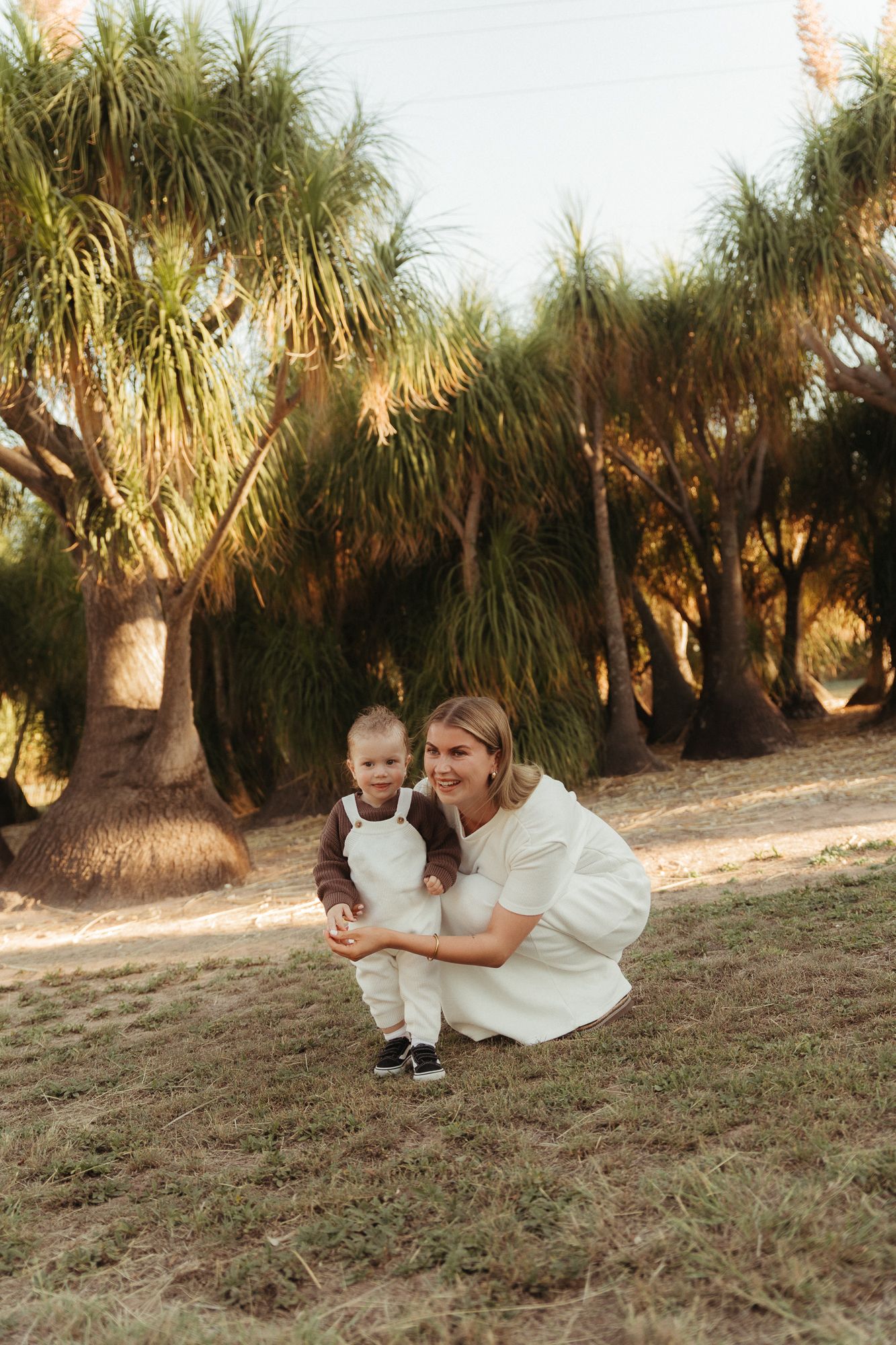 A smiling woman in a white dress crouches beside a happy toddler outdoors, surrounded by tall palm trees in warm sunlight.