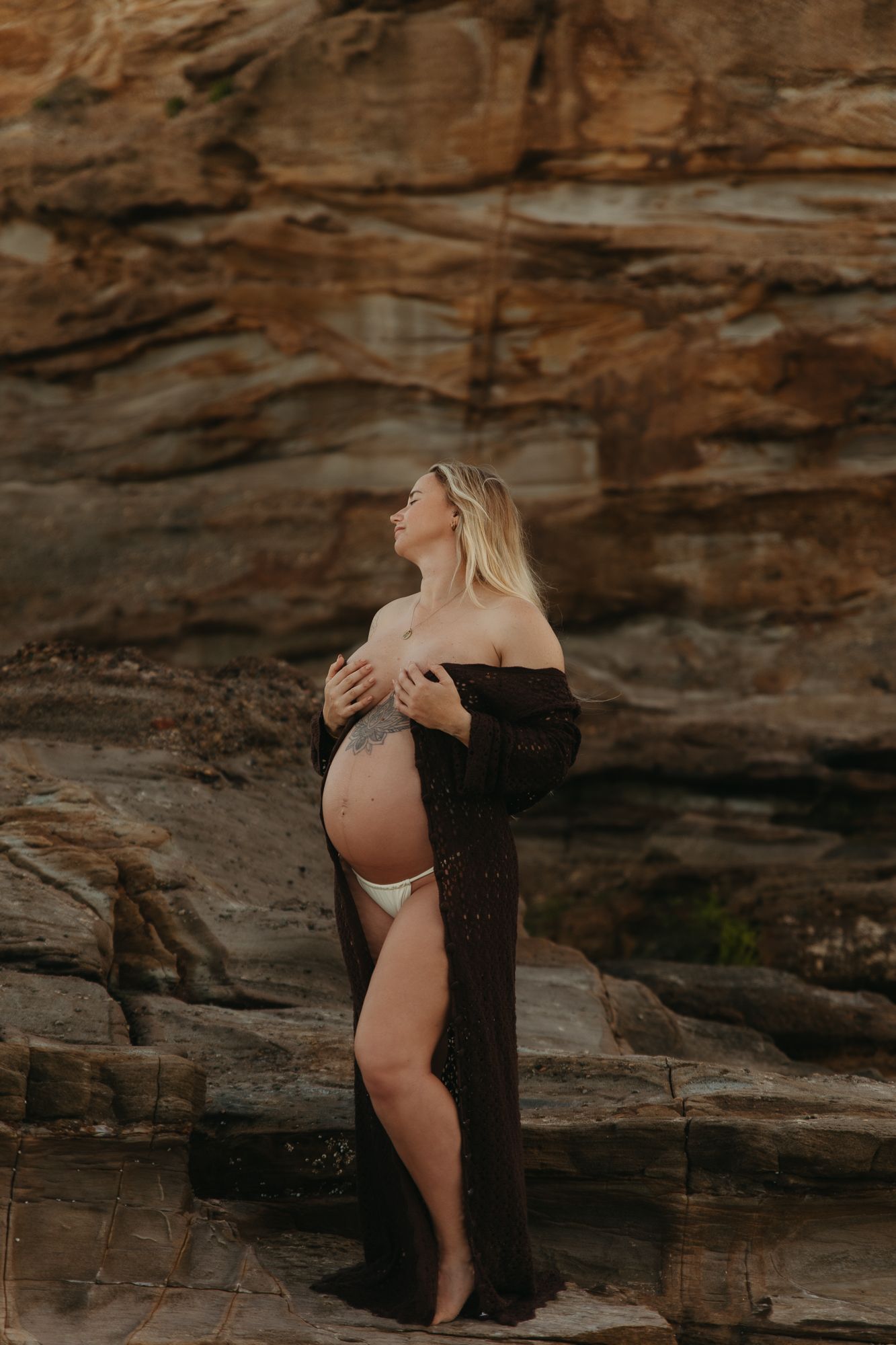Pregnant woman in dark robe poses against rocky cliff, cradling bare baby bump, wearing white bikini bottoms outdoors.