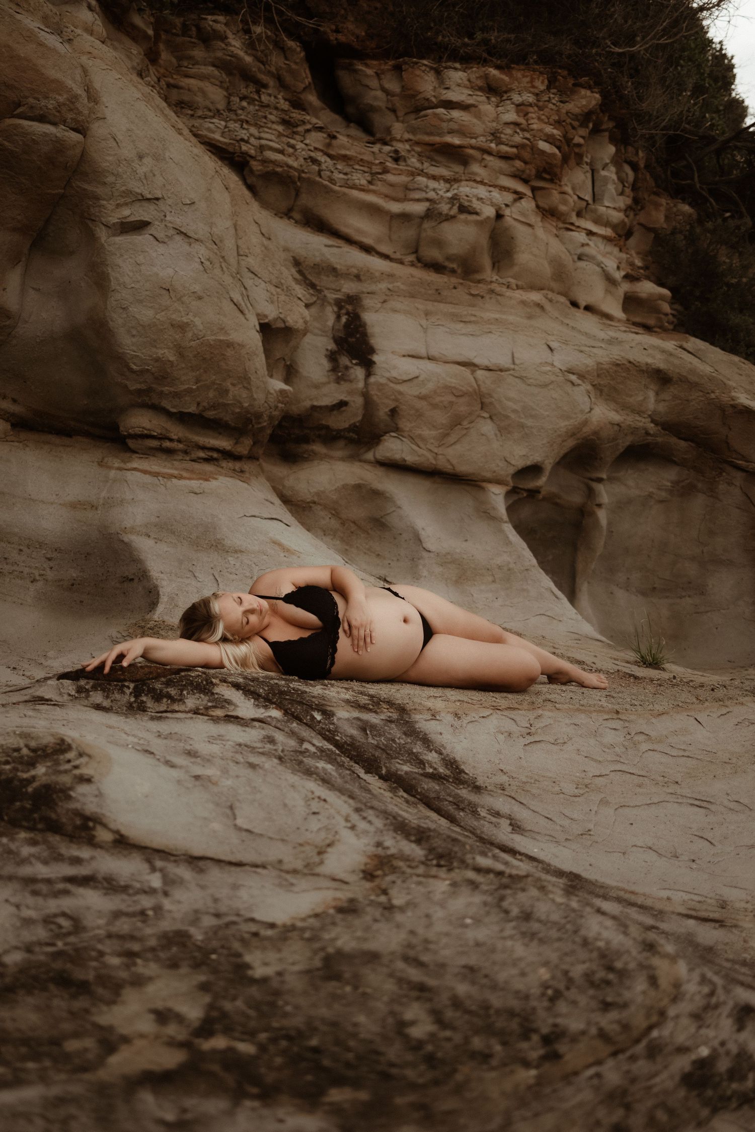 Pregnant woman in black bikini posing gracefully on smooth sandstone rocks near a cliff face.
