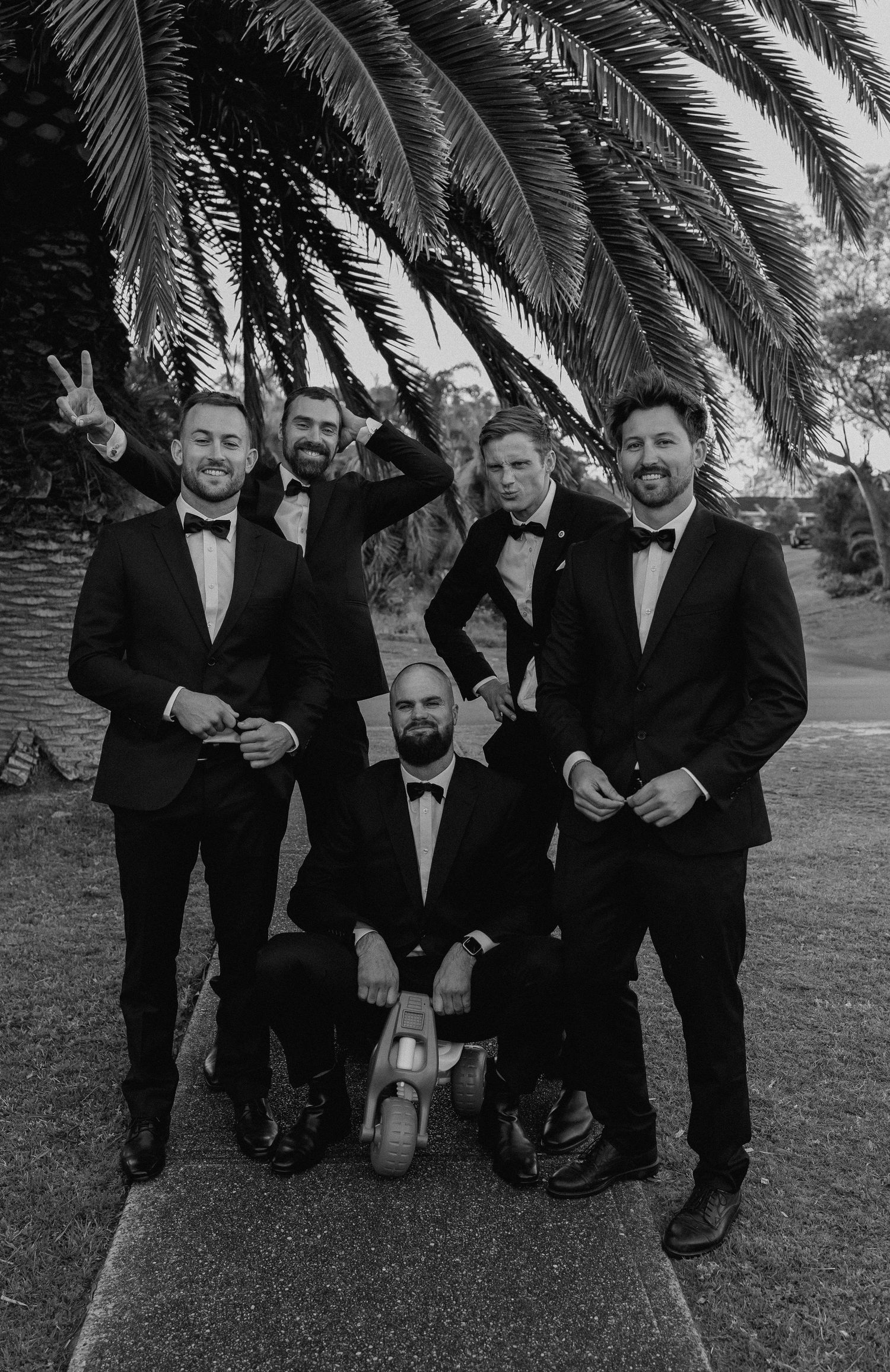 Black and white fun groomsmen photo posing under a palm tree in matching black tuxedos.