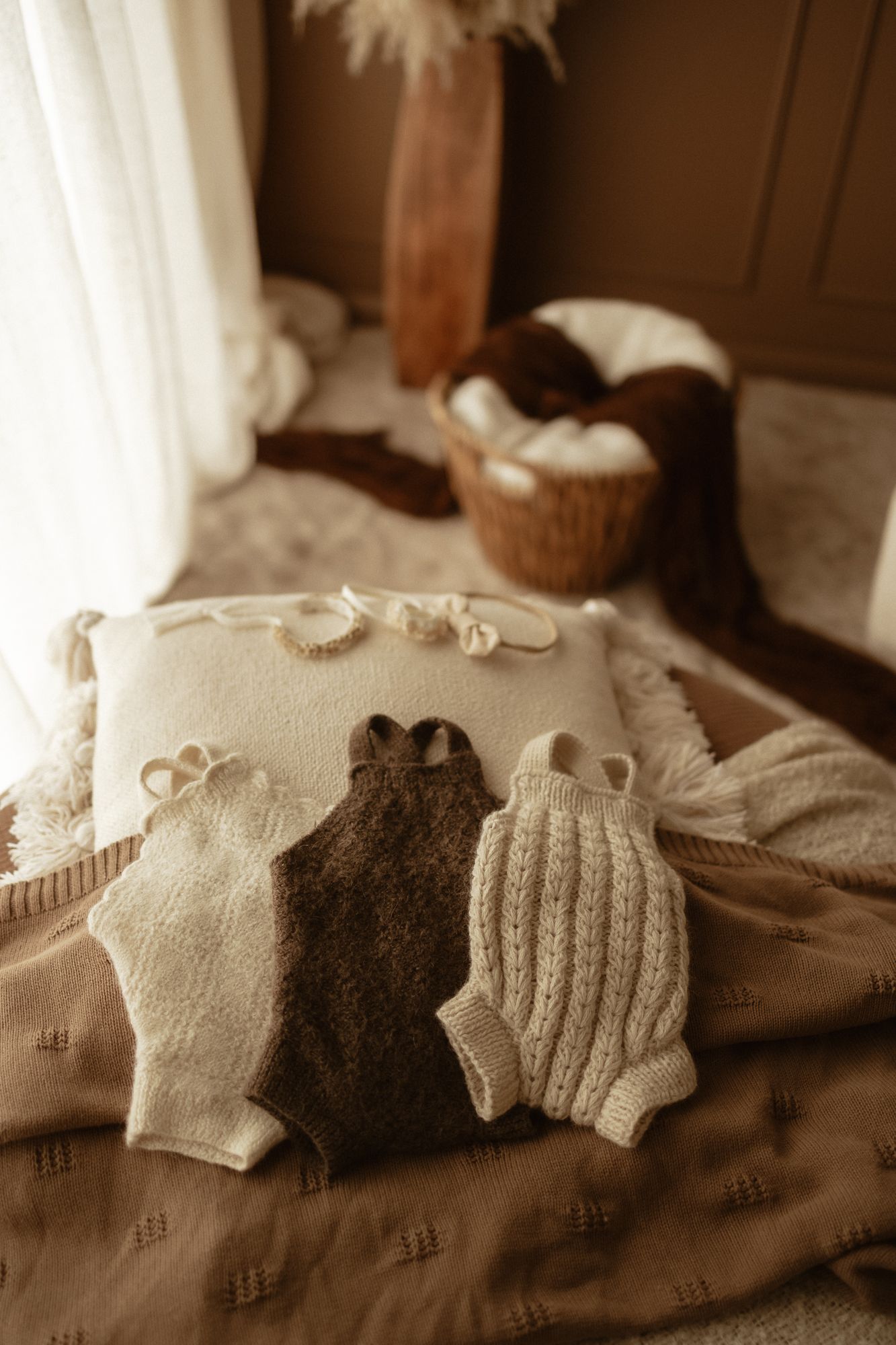 Three knitted baby rompers in cream and brown displayed on a decorative pillow with a cozy boho nursery setting.
