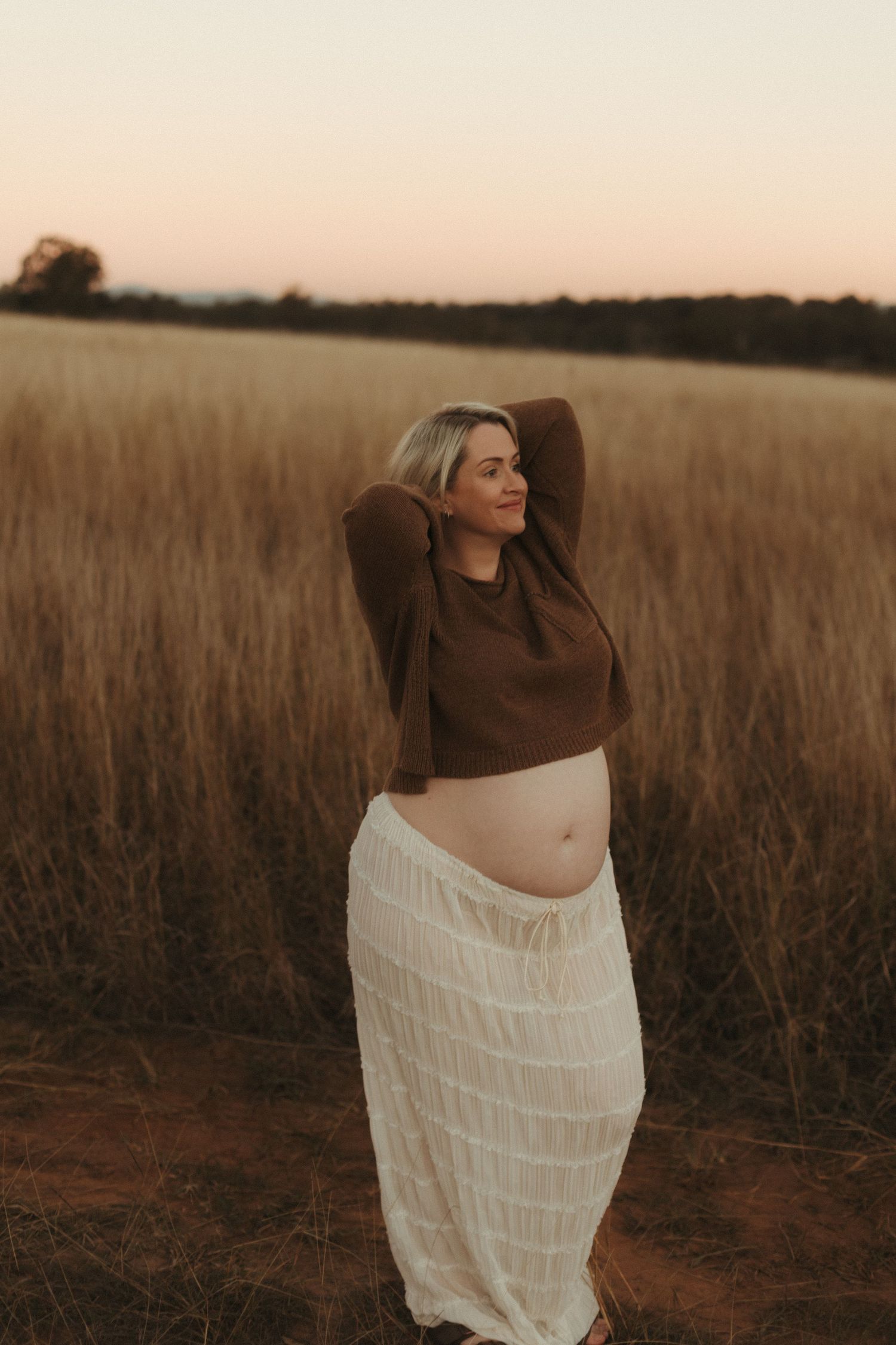 Smiling pregnant blonde woman lifting arms behind head in golden field at dusk.