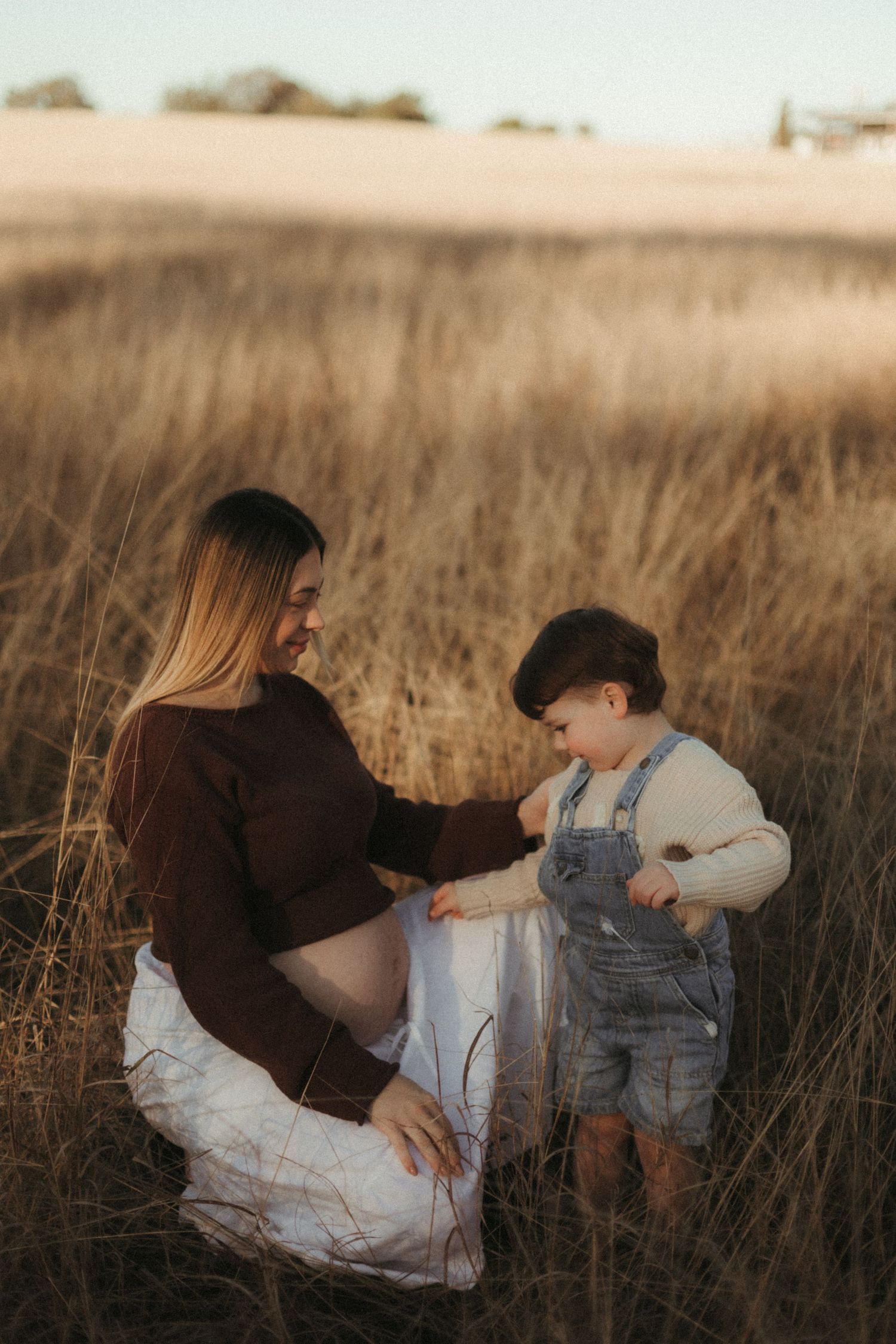 Smiling pregnant woman in brown top crouching beside toddler in denim overalls in a wheat field.