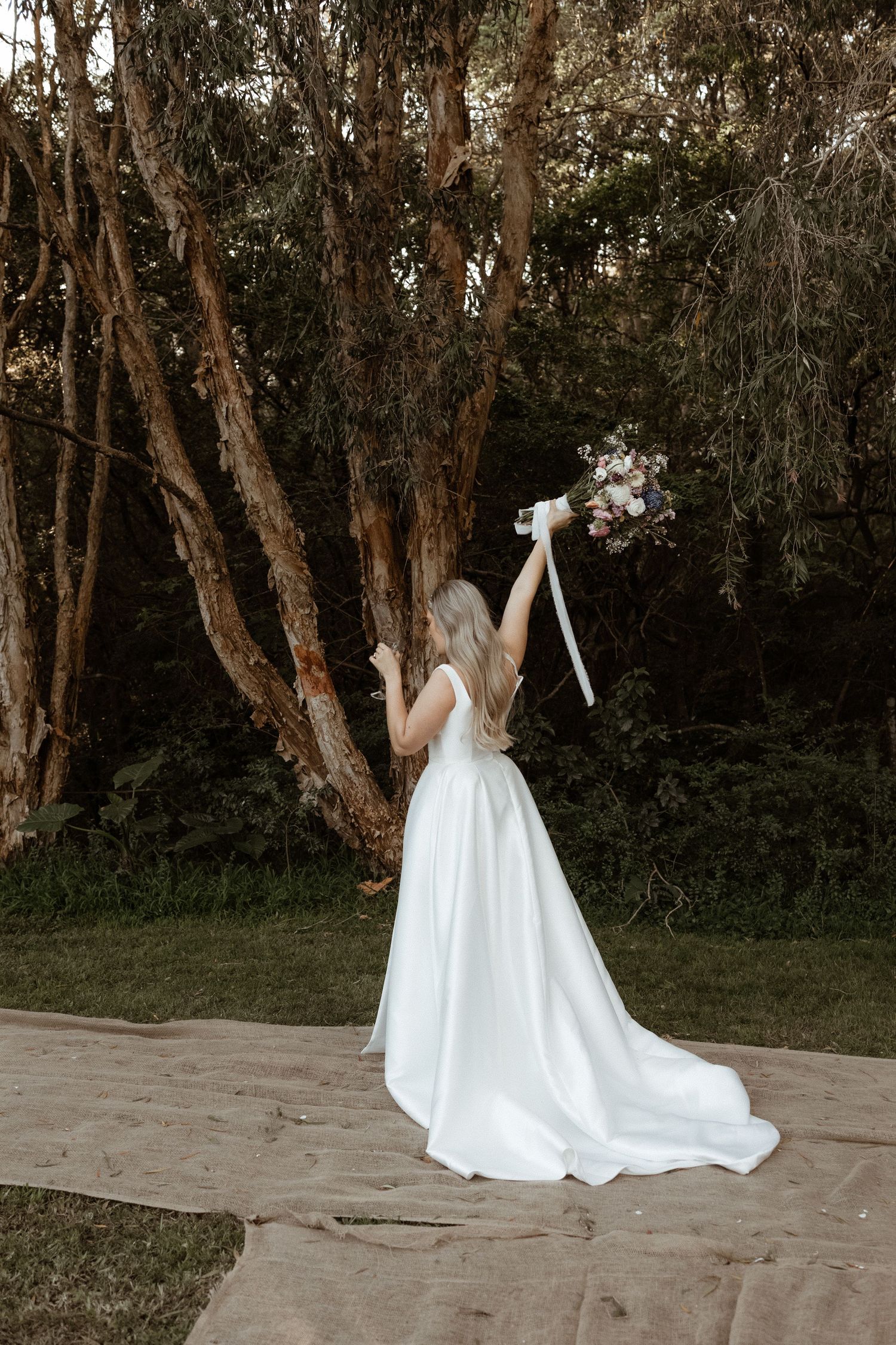 Bride in elegant white satin gown lifting bouquet with ribbon streamers among tall trees outdoors.