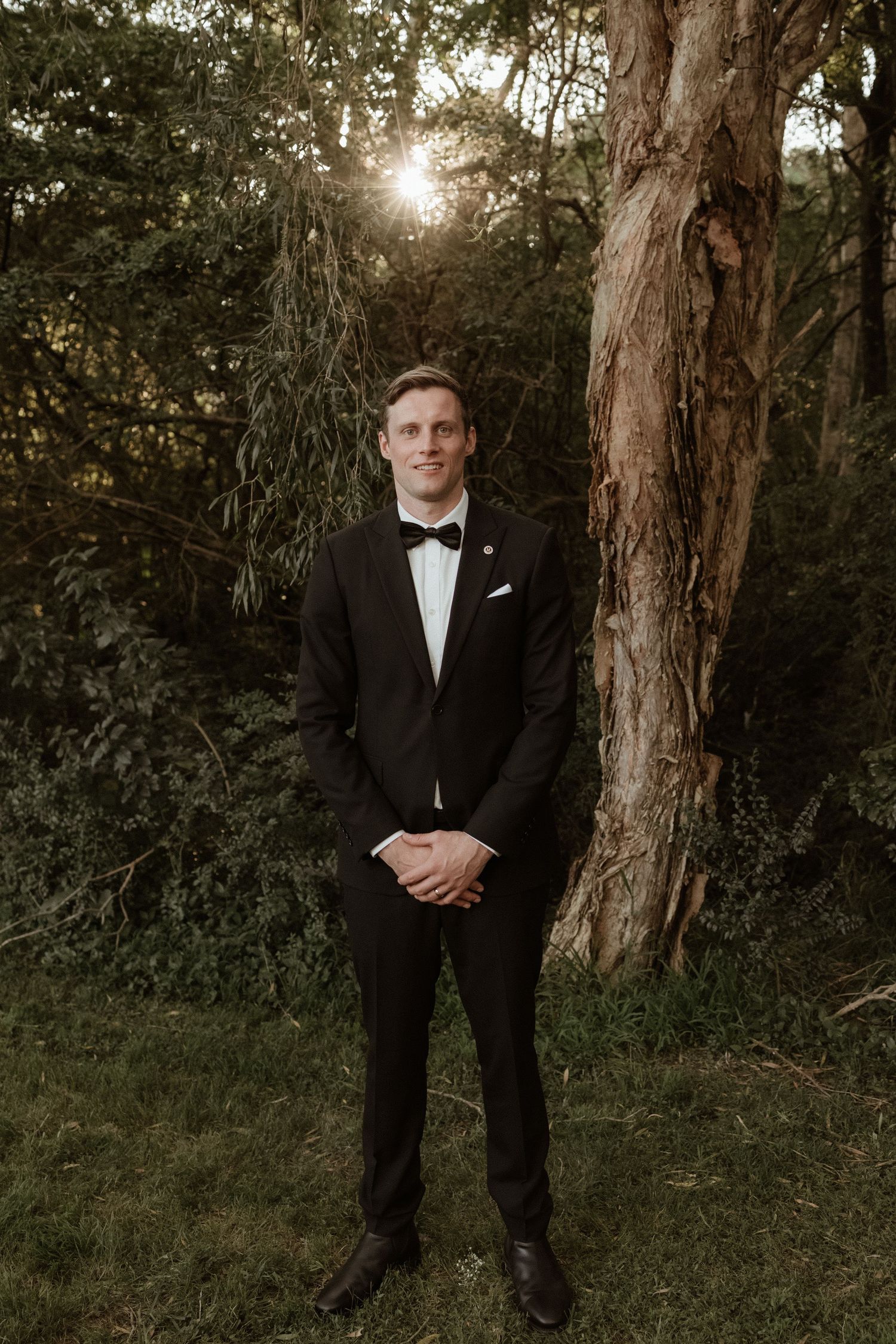 Smiling groom in black tuxedo and bow tie stands outdoors near a large tree with sunlight filtering through green foliage.