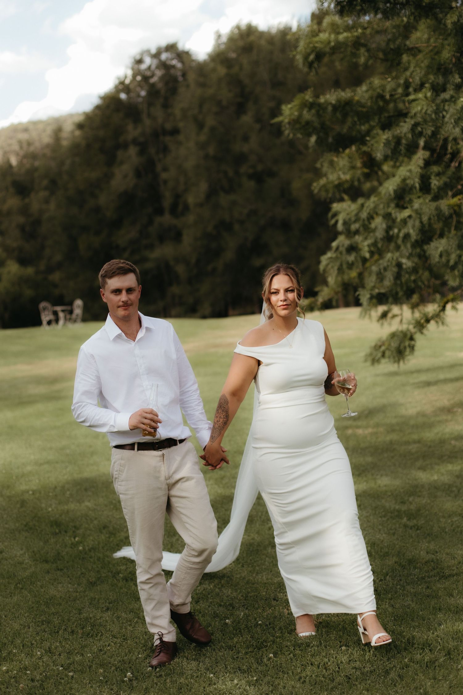A couple in white wedding attire holds hands while walking on a lush green lawn with trees in the background.