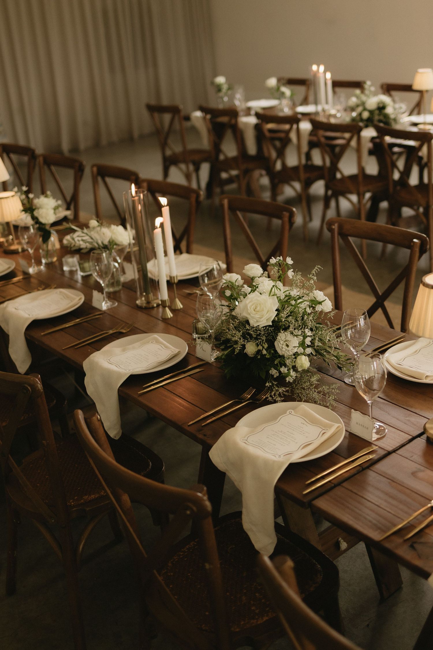 Elegant rustic wedding reception with dark wood farm tables, cross-back chairs, white floral centerpieces, and candlelight.