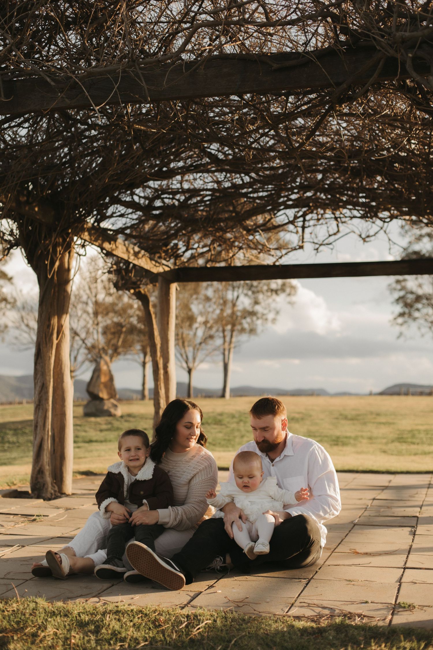 Family of four sitting on stone patio under bare pergola in golden light.