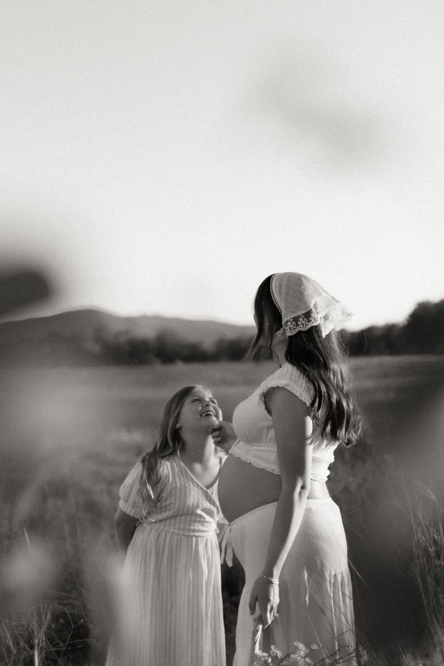 Black and white maternity photo of pregnant woman with young girl laughing in field.