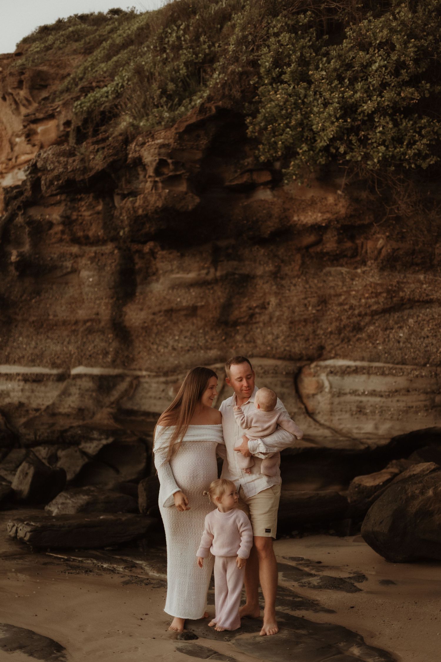 Pregnant family in white and pink outfits posing against sandstone cliffs on beach.