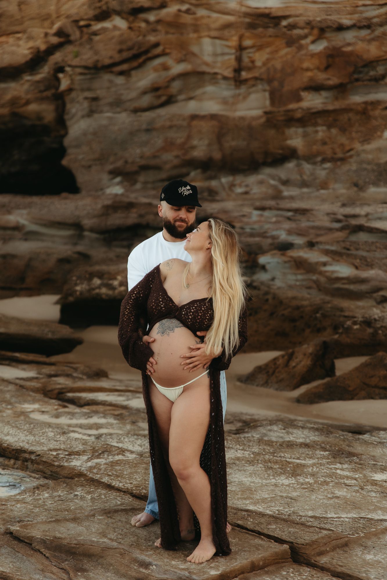 Couple embracing on rocky beach, pregnant woman in lace robe and white bikini, man in black cap and white shirt.