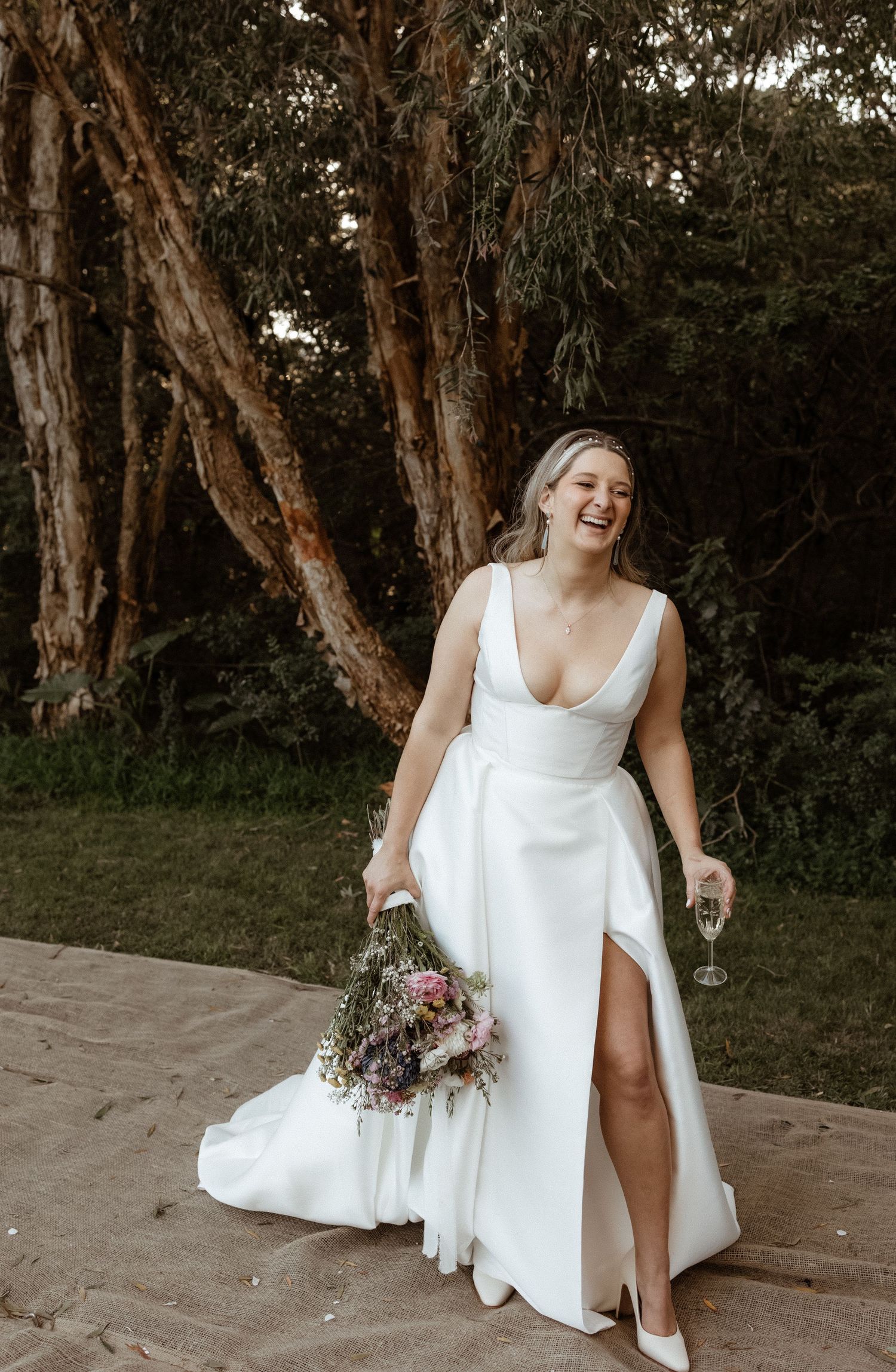 Laughing bride in white gown with slit holds bouquet and champagne outdoors near trees.
