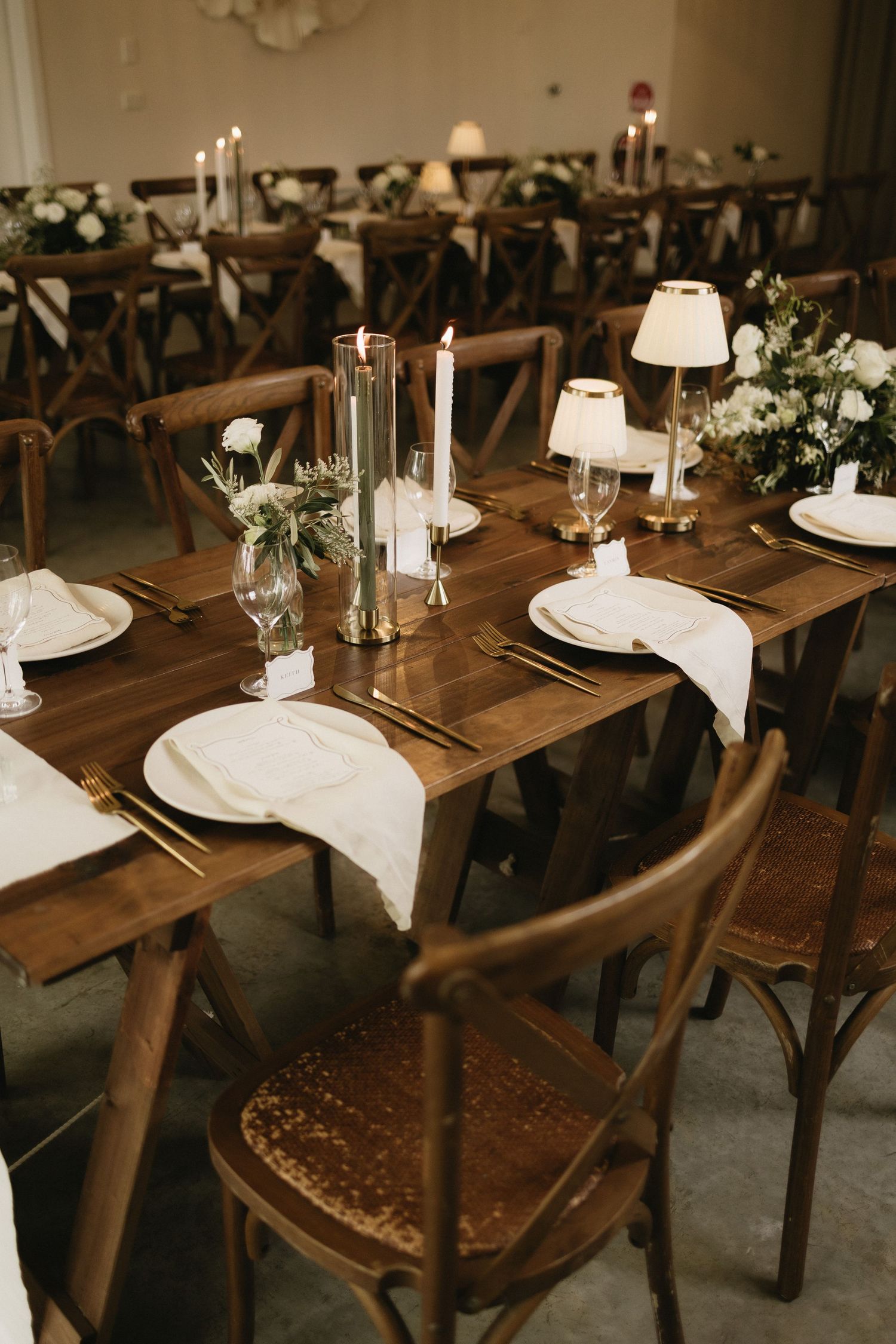 Elegant wedding reception tables with wooden cross-back chairs, white linens, candles, and floral centerpieces.