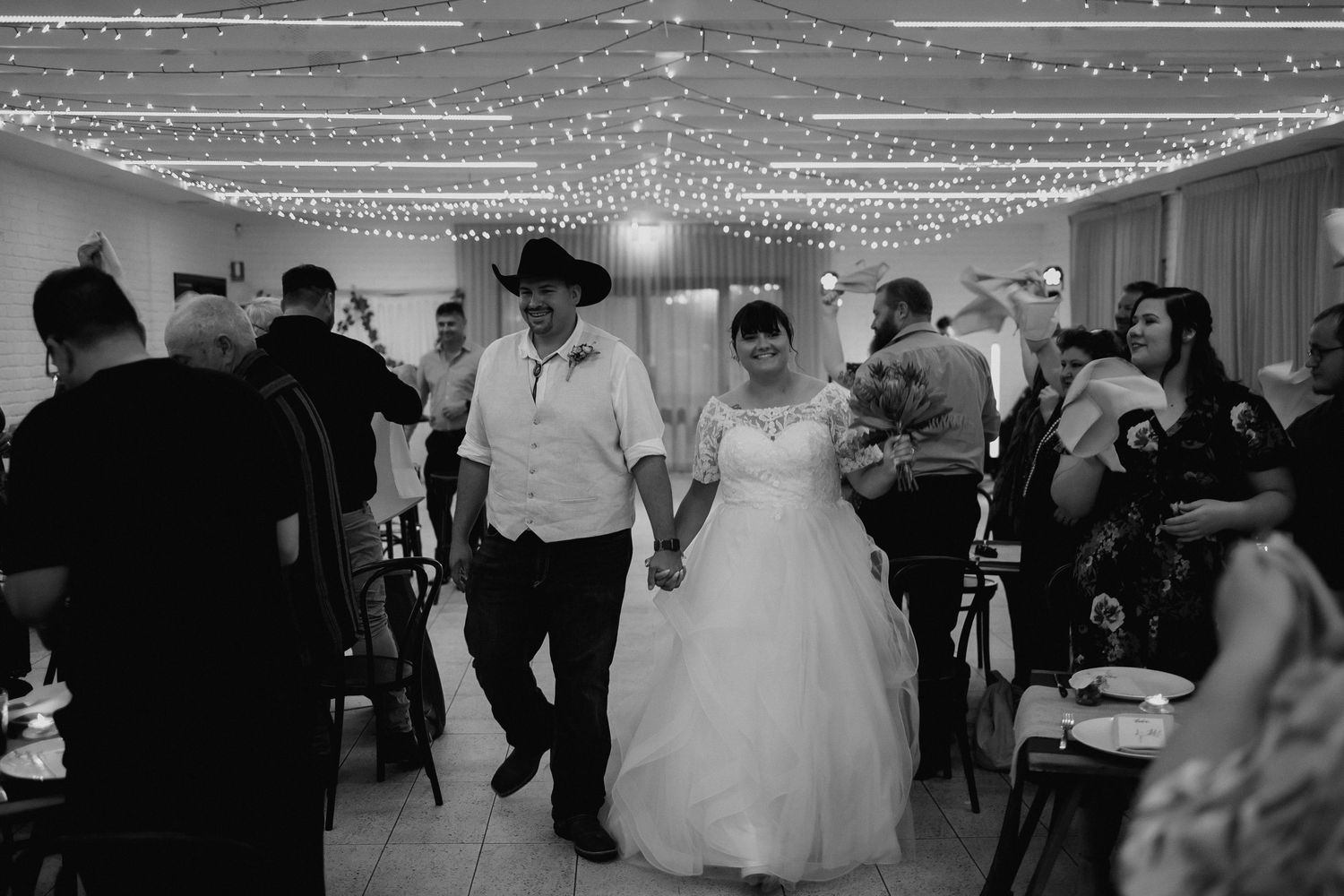 Bride and groom walking hand in hand into wedding reception under fairy lights.