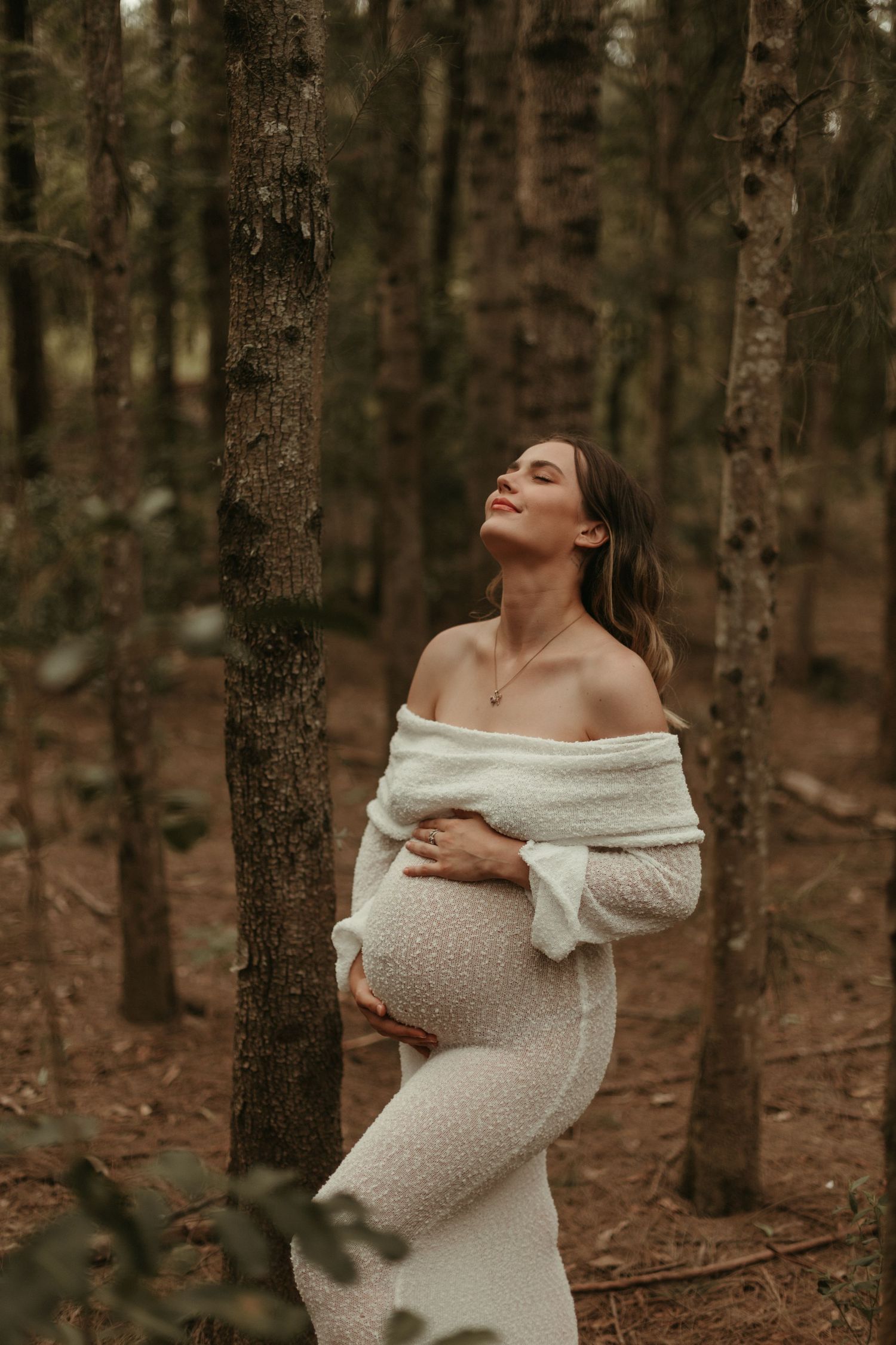 Pregnant woman in off-shoulder white knit dress stands among forest trees, eyes closed, holding her bump.