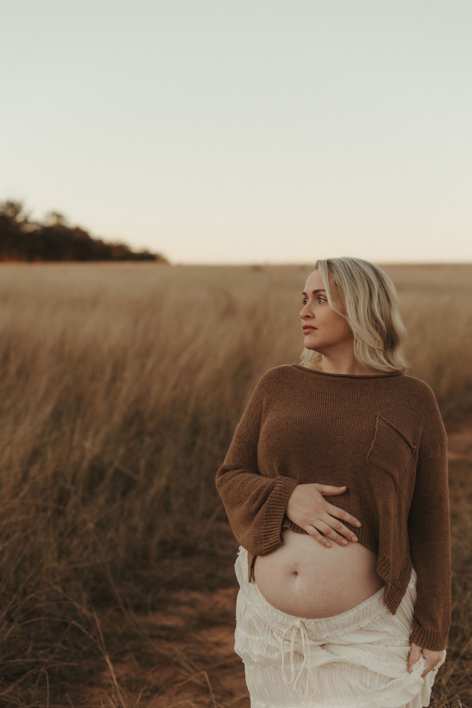 Pregnant woman in brown knit sweater cradling baby bump in golden wheat field at sunset.