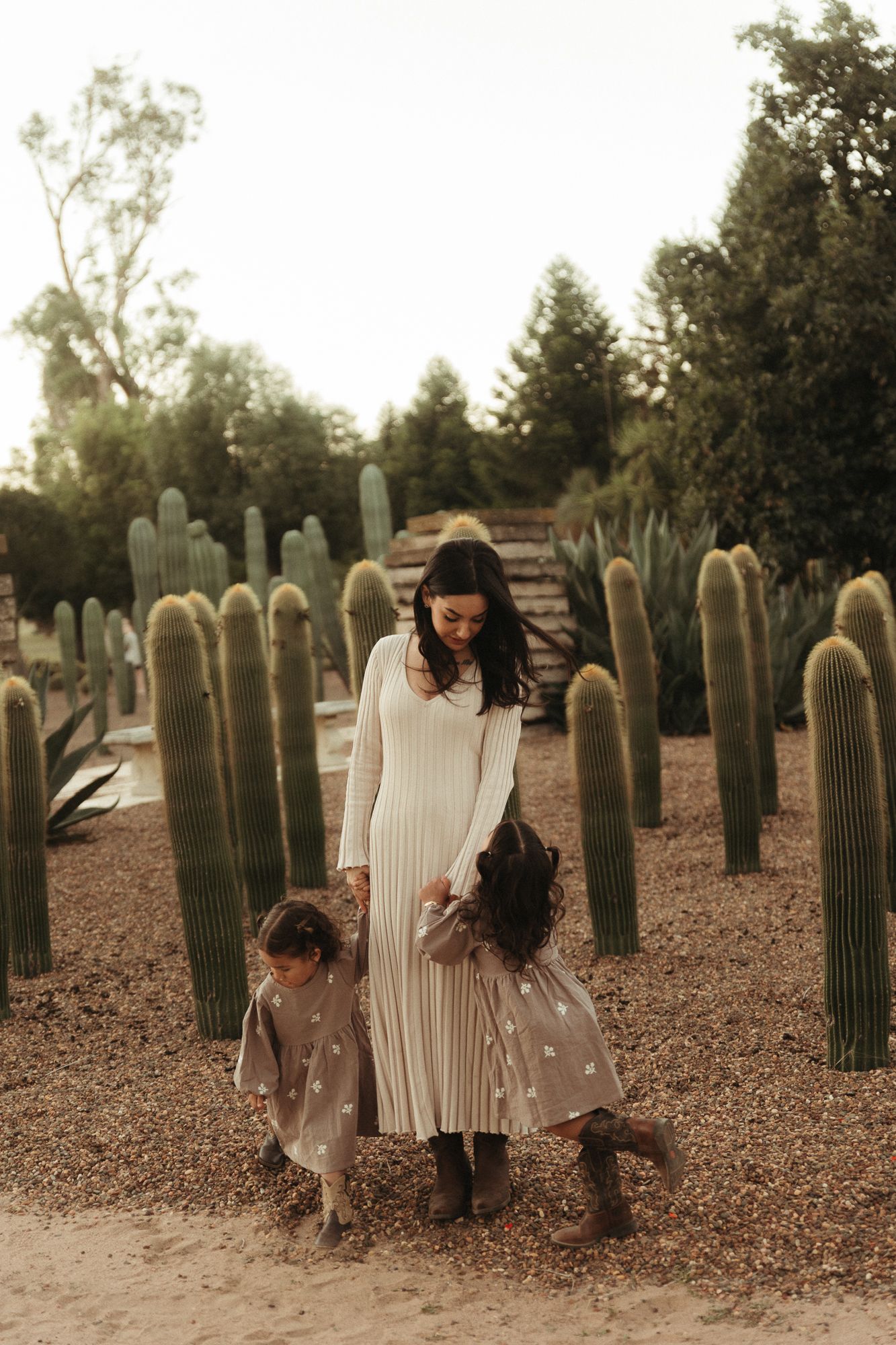 A woman in a white dress walks with two young girls in matching outfits among tall cacti in a desert garden.