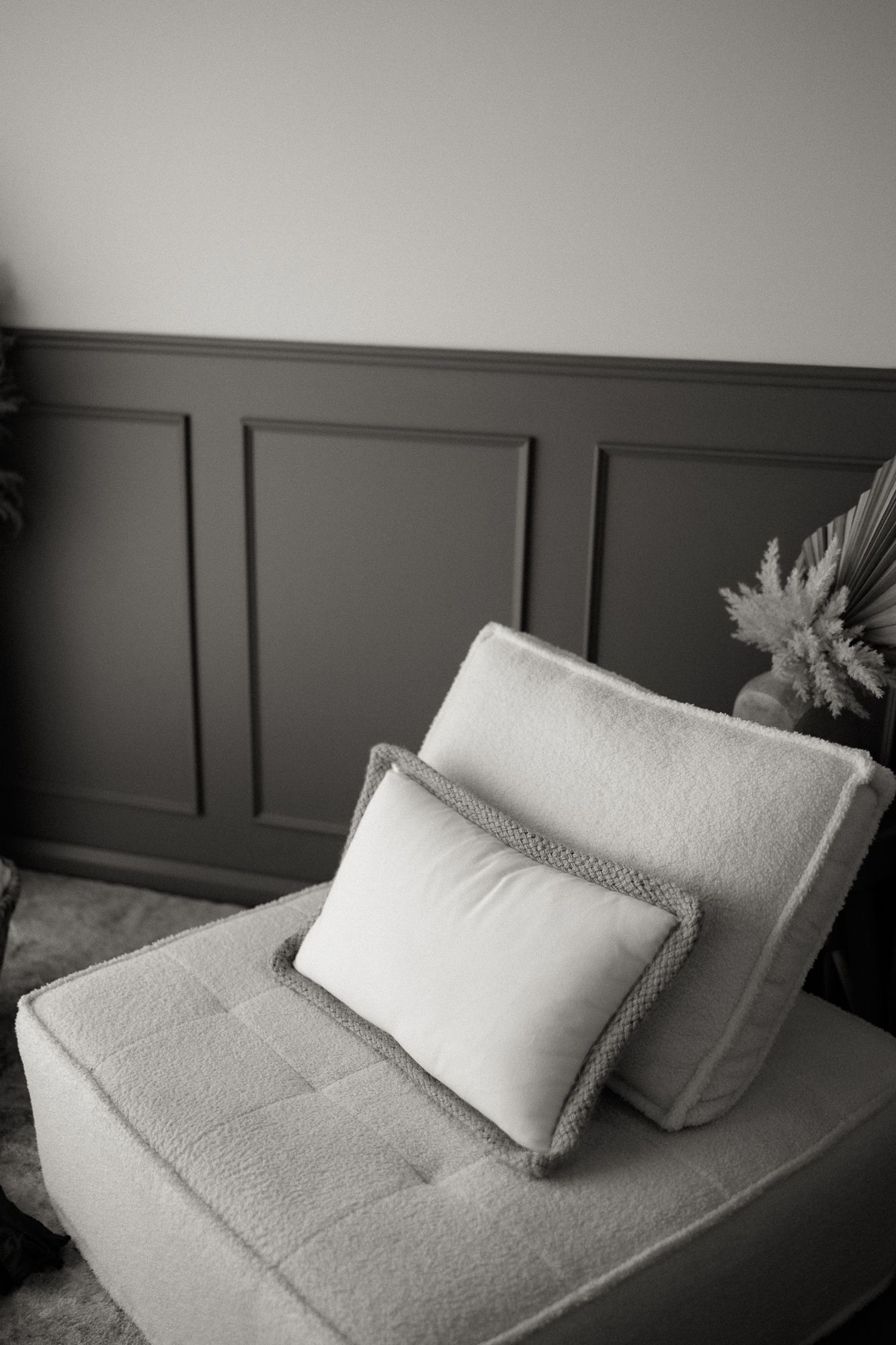 A white boucle armchair with a decorative fringed pillow against a dark grey paneled wall.