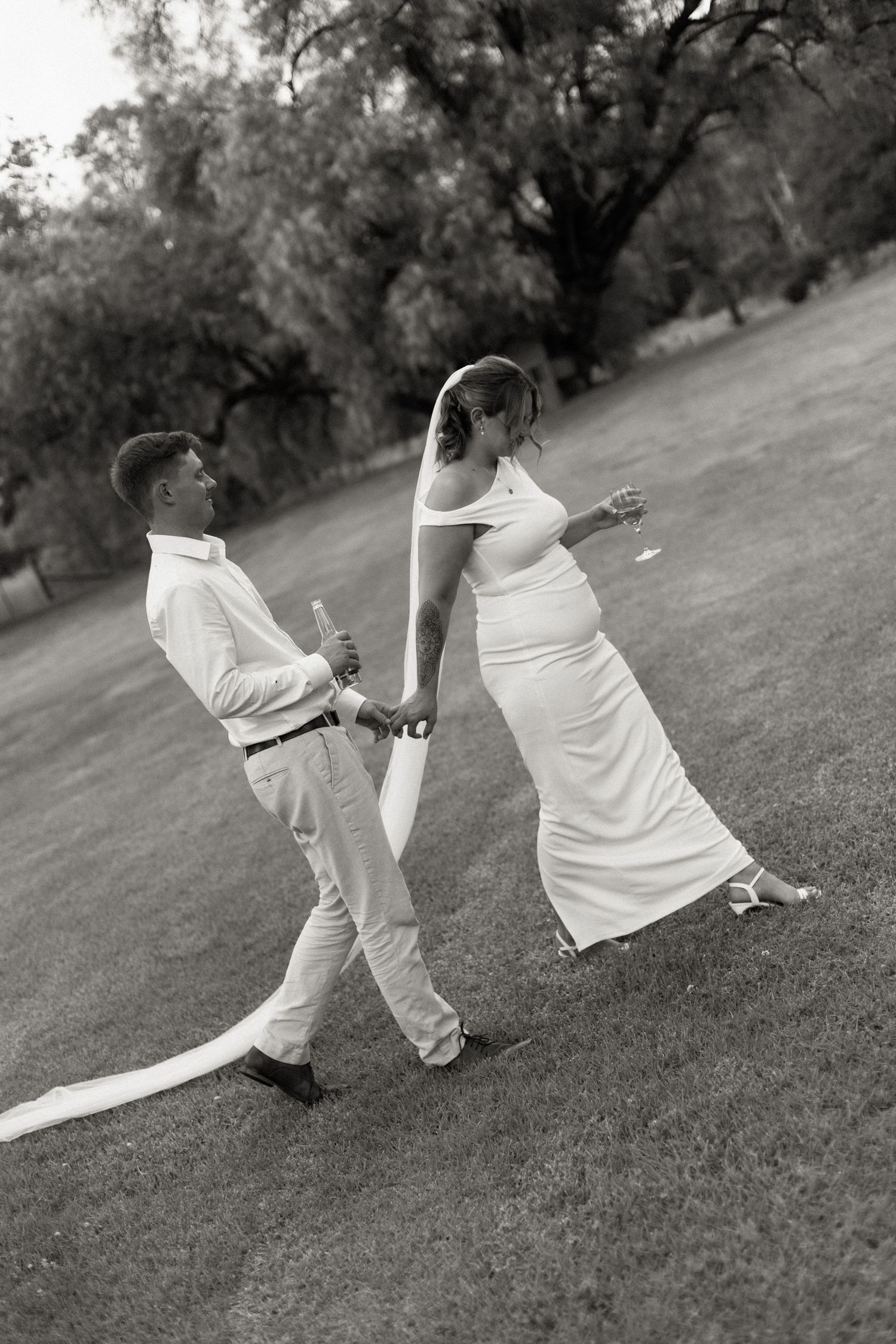 Black and white photo of newlyweds strolling across green lawn with flowing bridal veil.