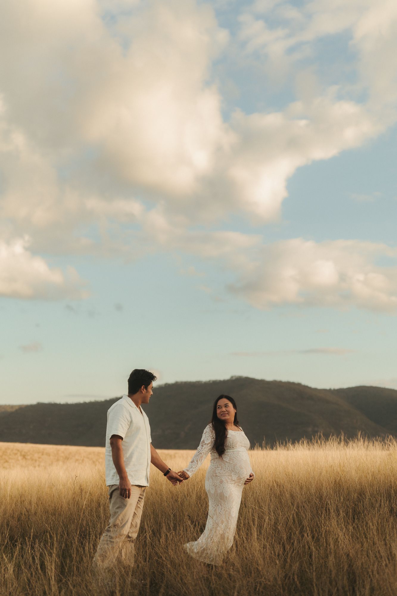 Couple holding hands in golden wheat field, pregnant woman in white lace dress, mountains and cloudy sky backdrop.