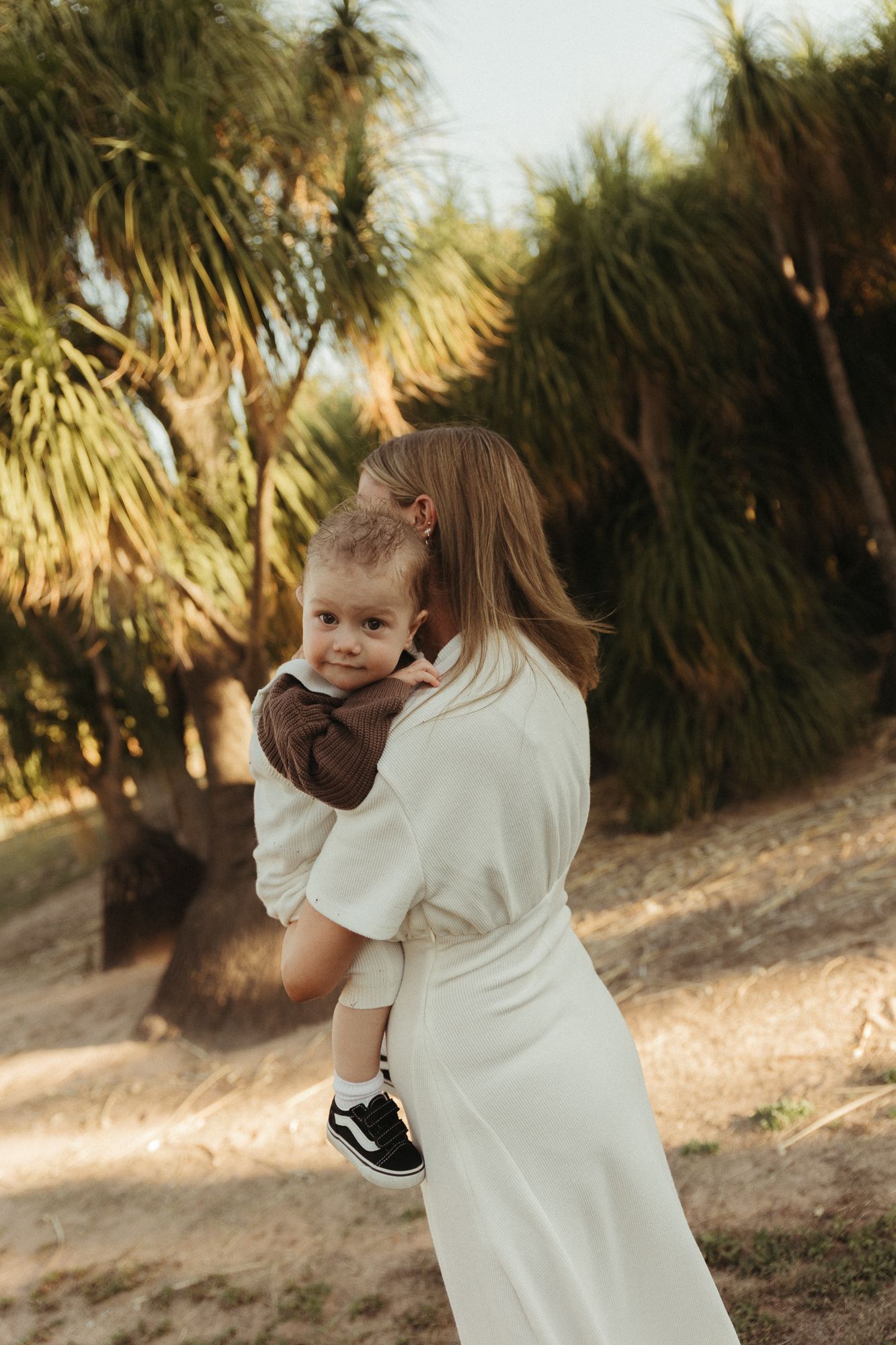 A mother in a white dress holds her toddler outdoors near palm trees in warm golden sunlight.