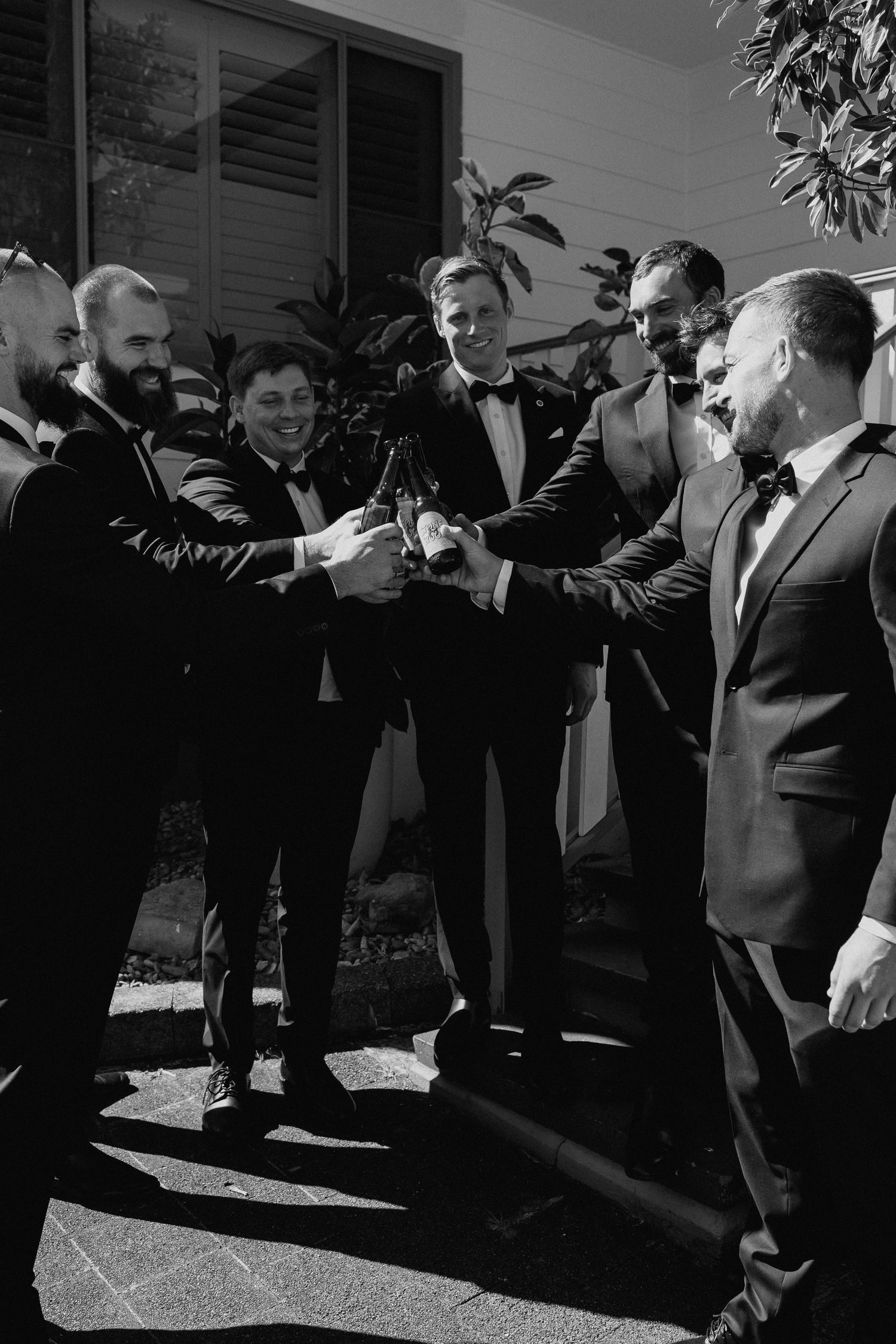 Black and white photo of groomsmen toasting with beer bottles outdoors.