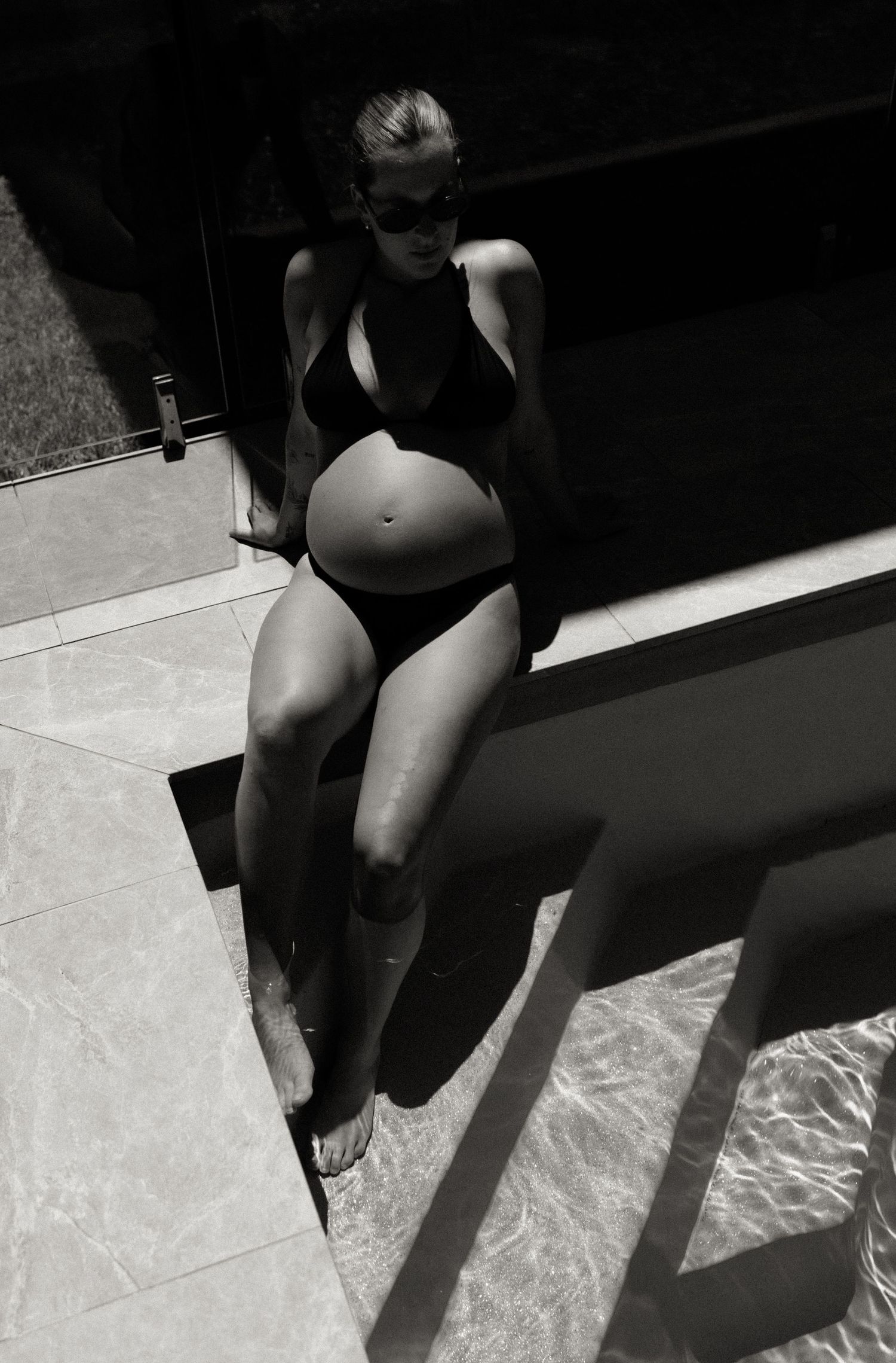 Black and white photo of a pregnant woman in a black swimsuit sitting by a sunlit pool.