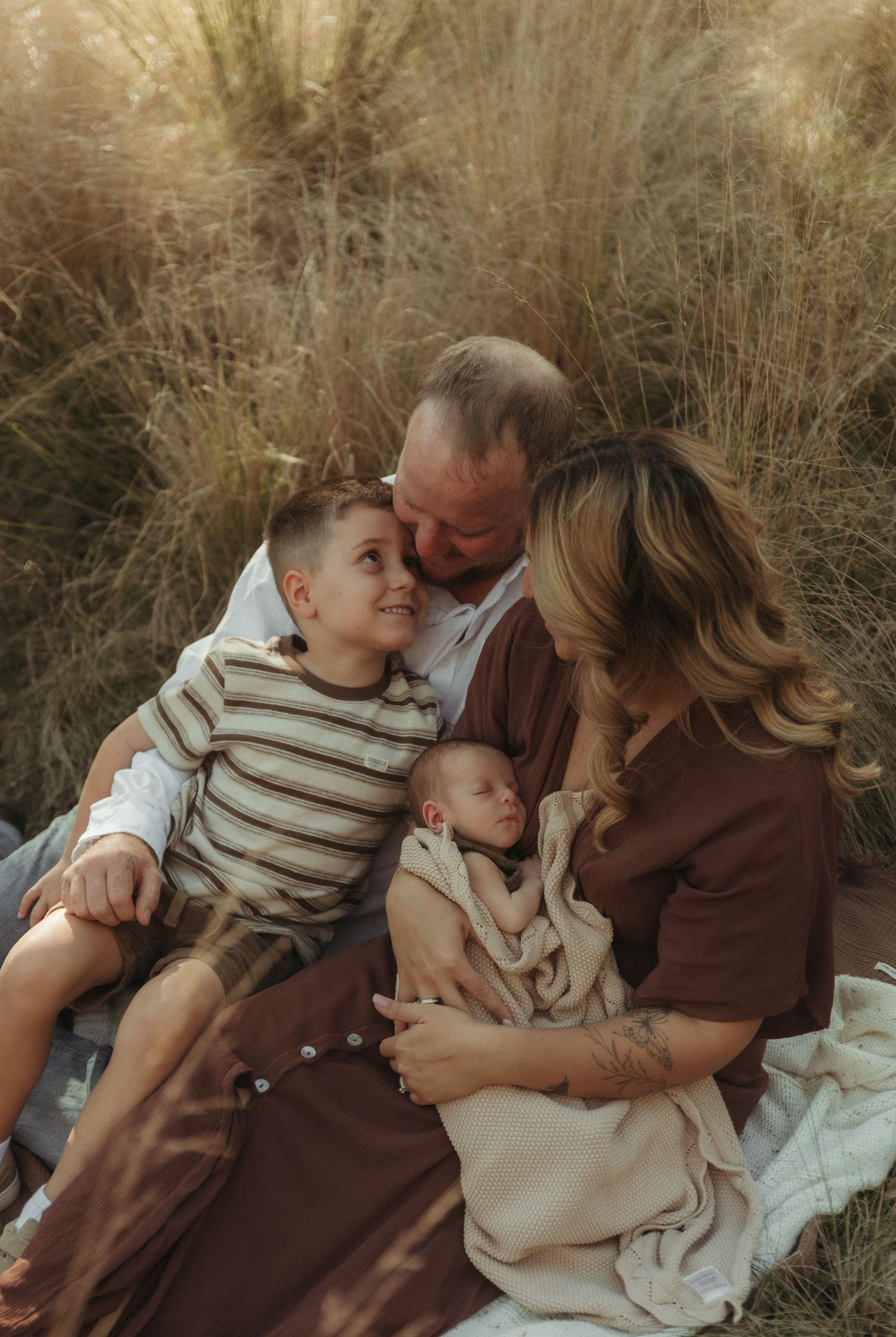 Family of four with newborn baby and young son cuddling together in a golden grass field.