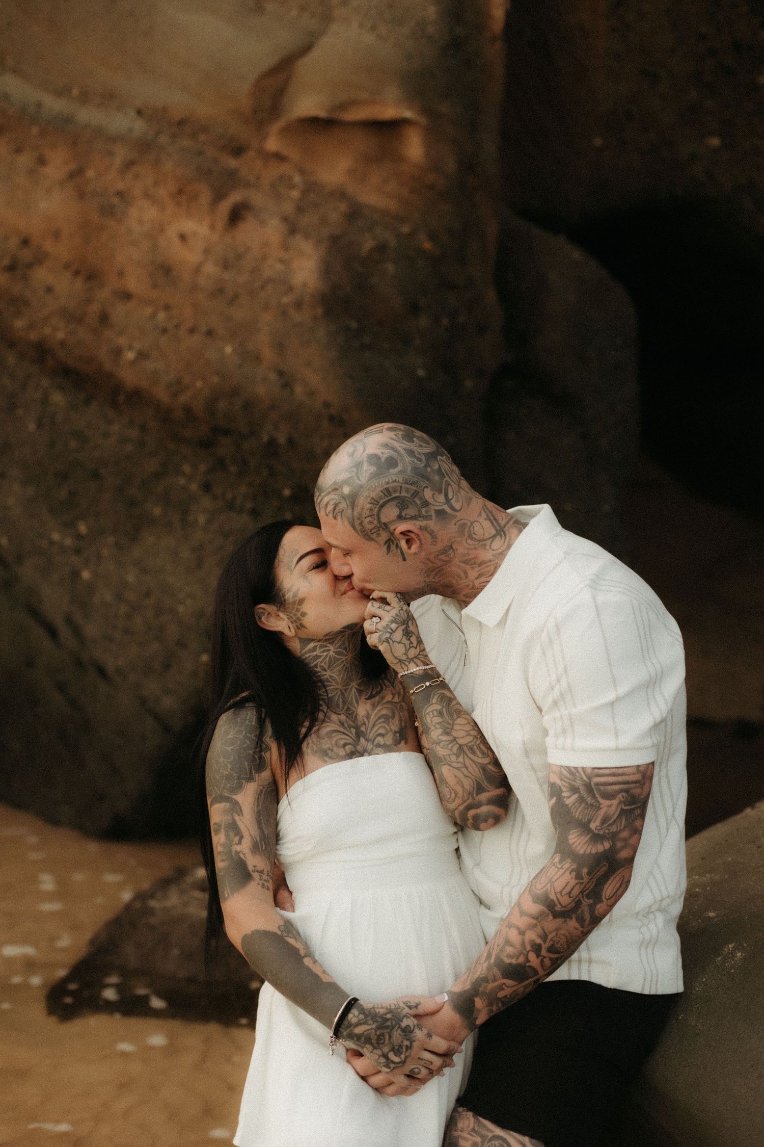 Heavily tattooed couple kissing in front of large rocks, woman in white maternity dress.