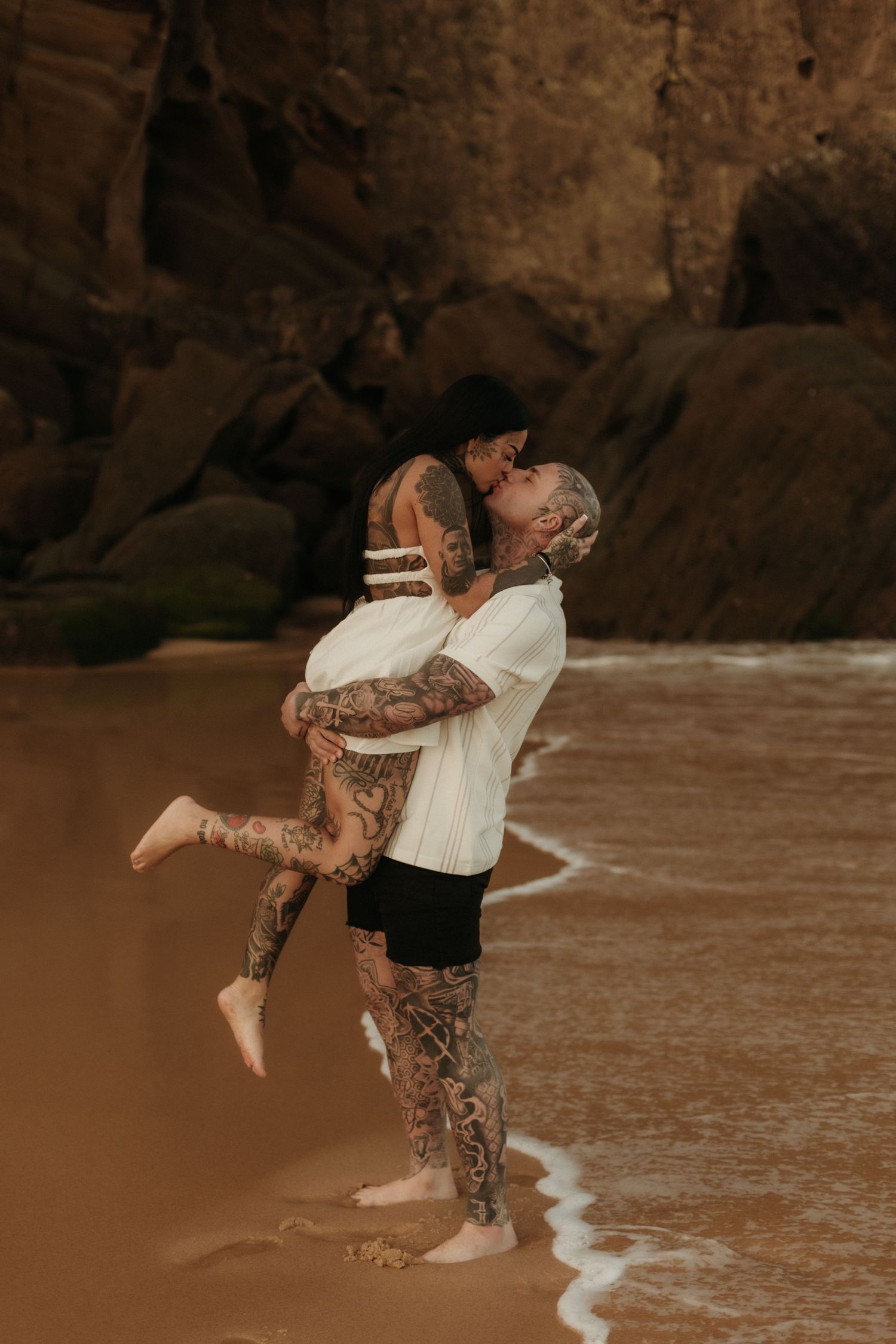 Tattooed man lifting woman in his arms on rocky beach with ocean waves.