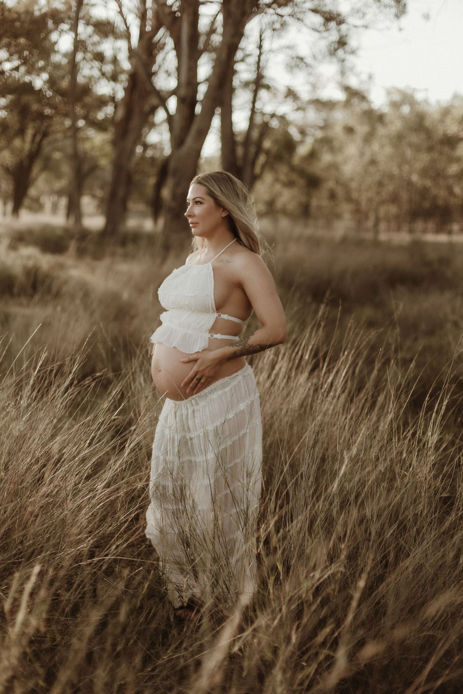 Pregnant woman in white halter maternity dress standing in tall golden grass among trees at dusk.