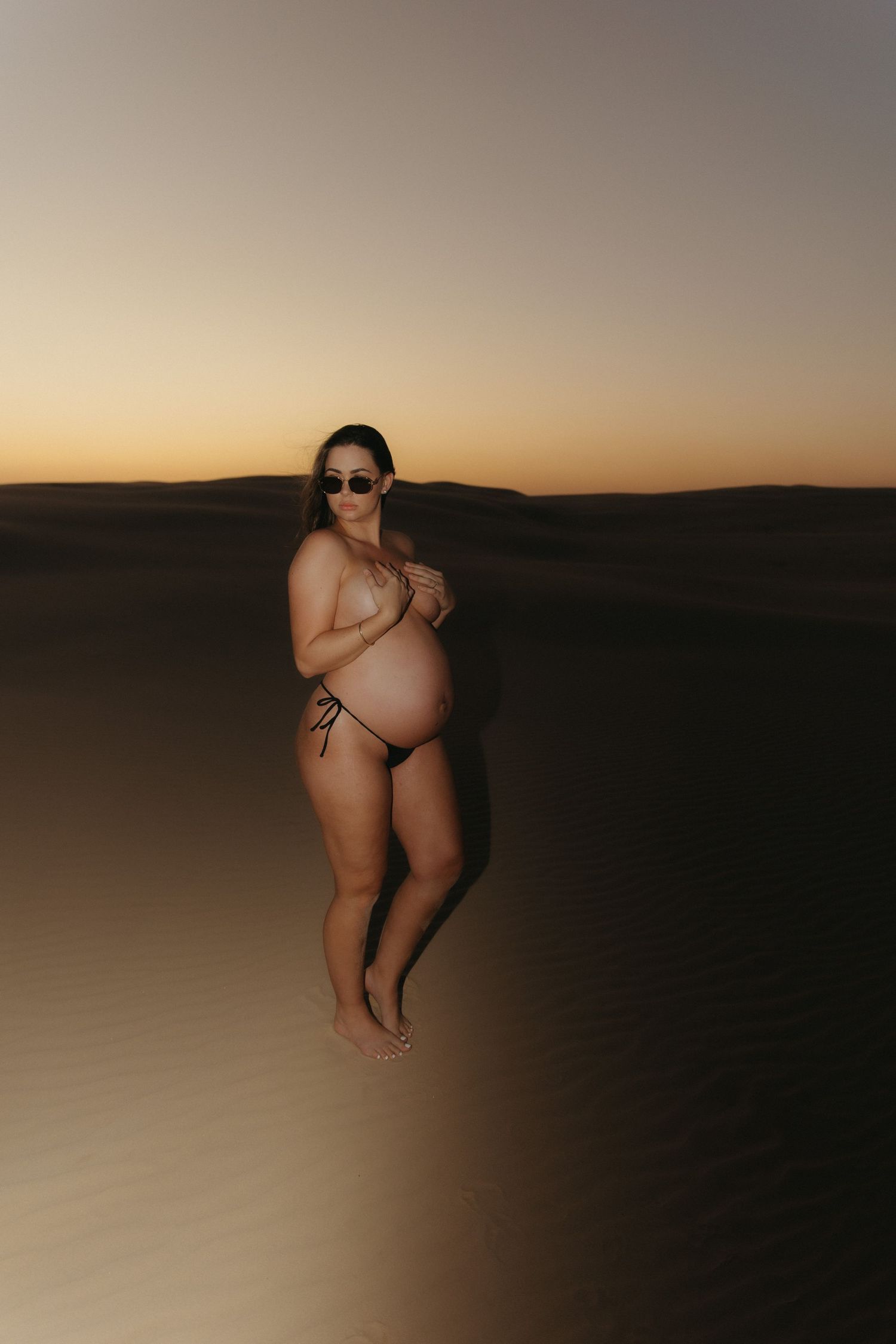 Pregnant woman in bikini and sunglasses posing on sand dunes at sunset in a desert landscape.