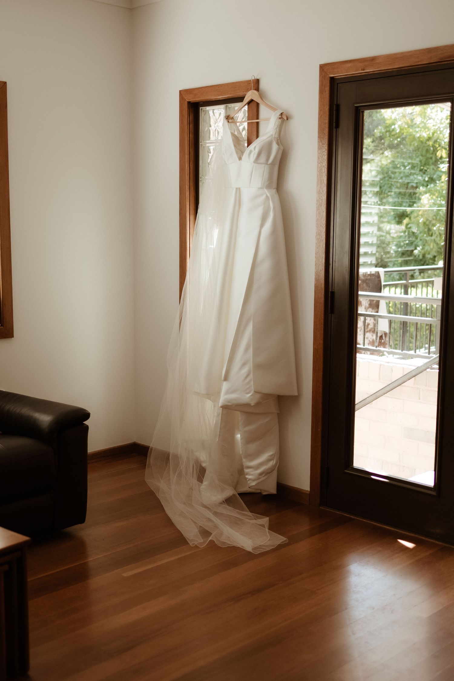 Elegant white wedding dress with veil hanging on a wooden door frame in a hotel room.