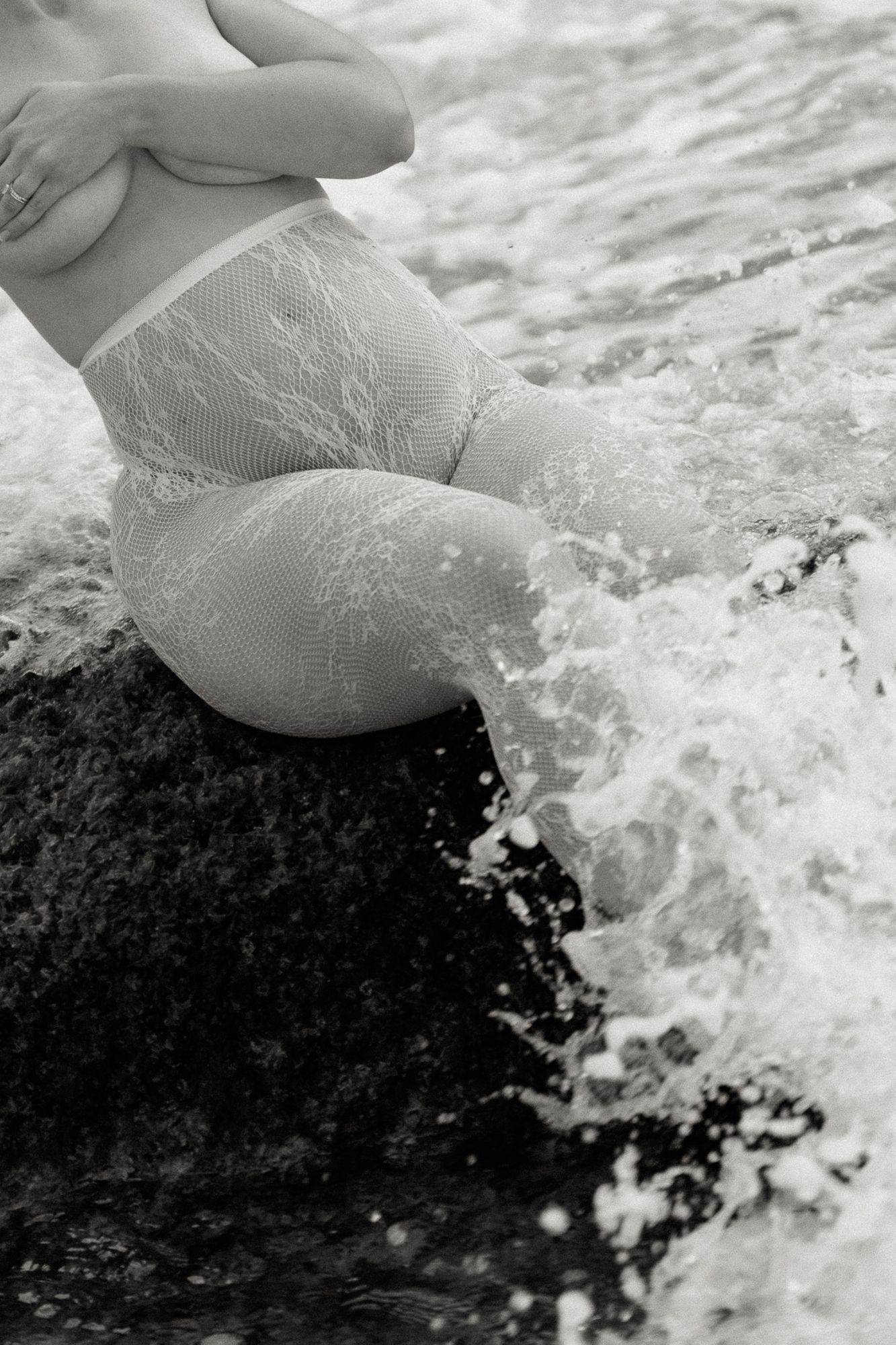 Black and white photo of a person in lace shorts sitting on a rock with ocean waves splashing around them.