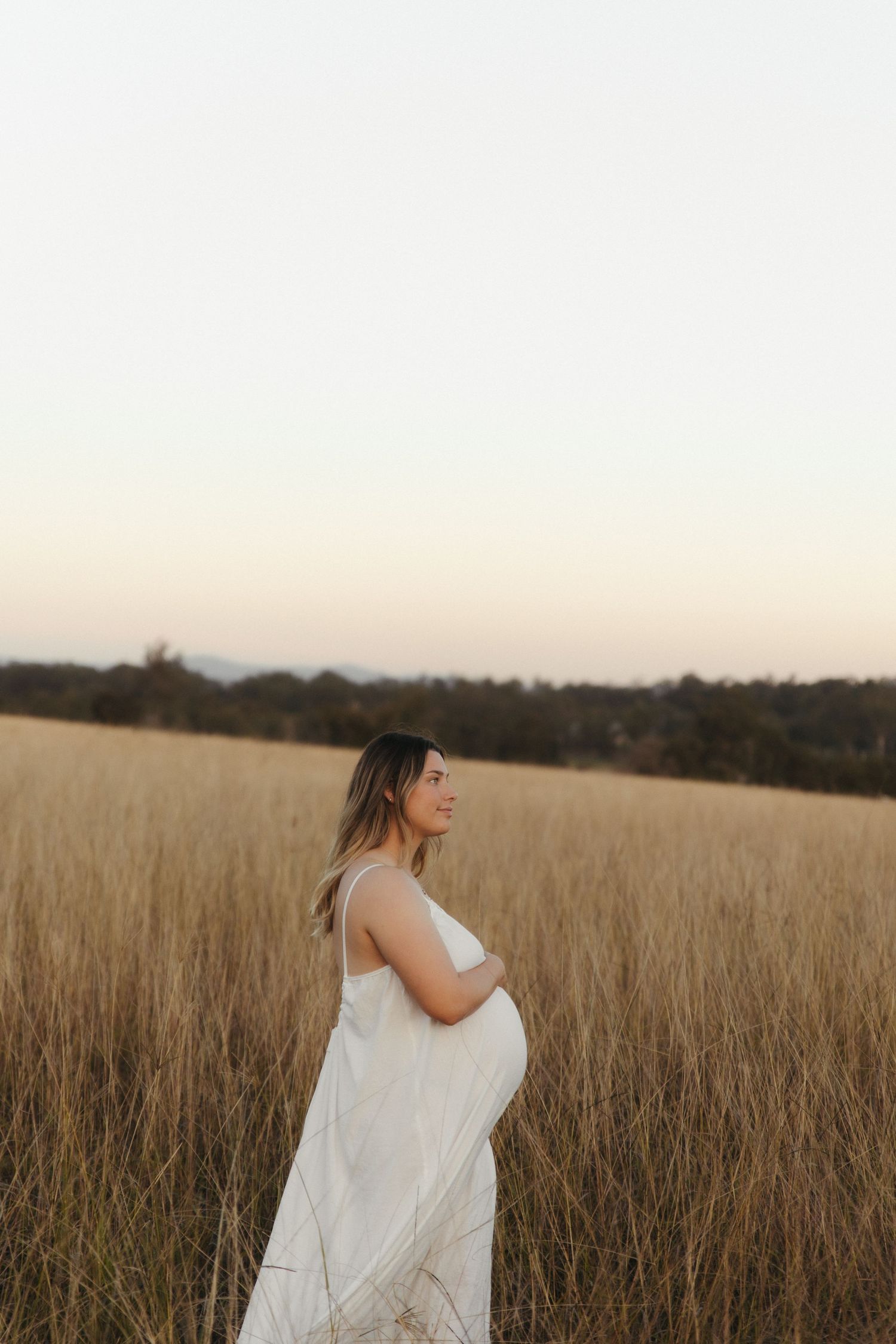 Pregnant woman in white dress standing in golden grass field at sunset, cradling her baby bump.