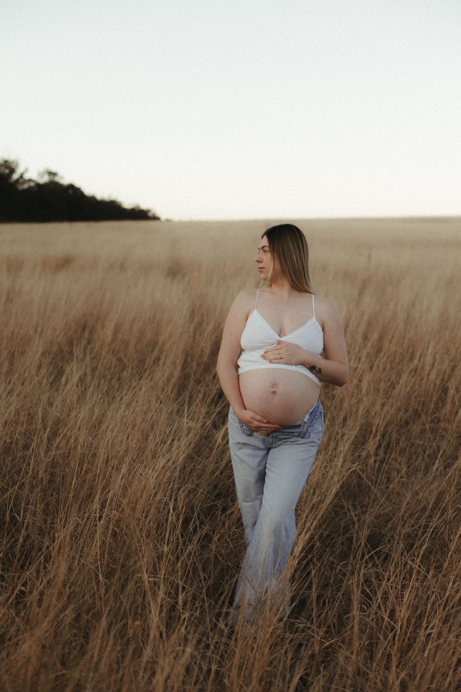 Pregnant woman in white bralette and jeans cradling her baby bump in a golden wheat field.